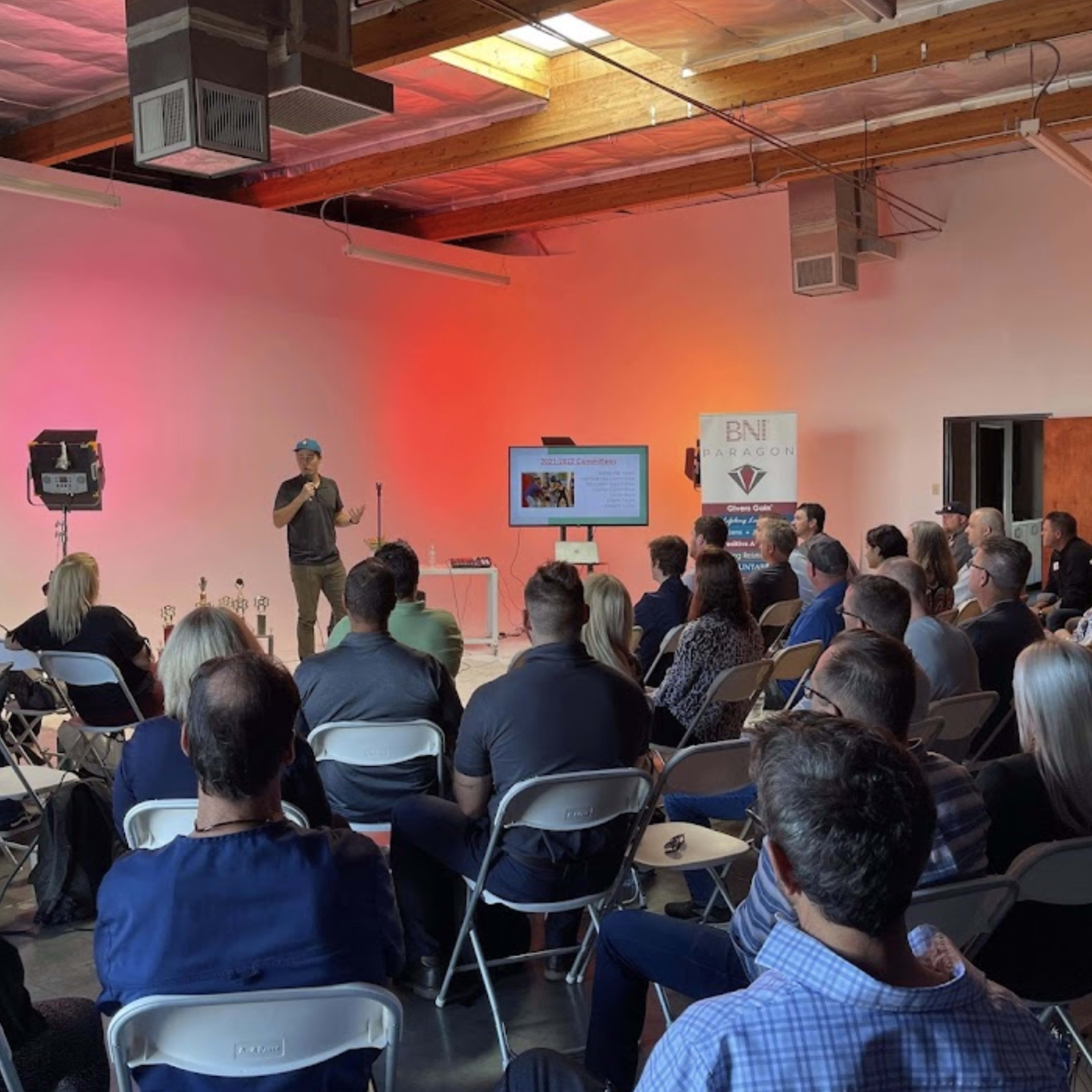 A speaker presents to an audience seated in a spacious venue with minimal decorations and a projector screen displaying a presentation.