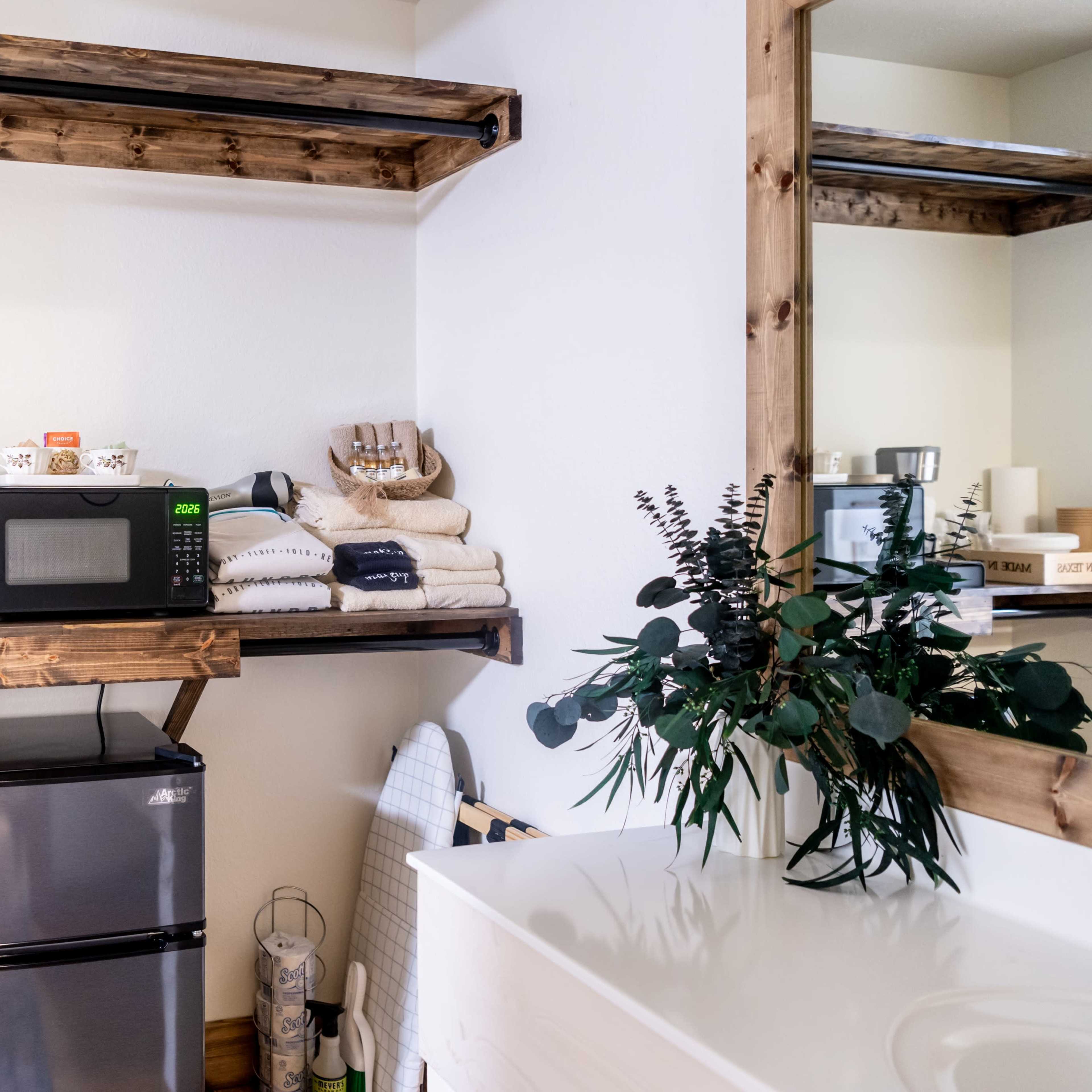 A shelf with a coffee maker, a microwave, neatly stacked towels, and a small refrigerator, with a bathroom vanity and a decorative plant in the background.