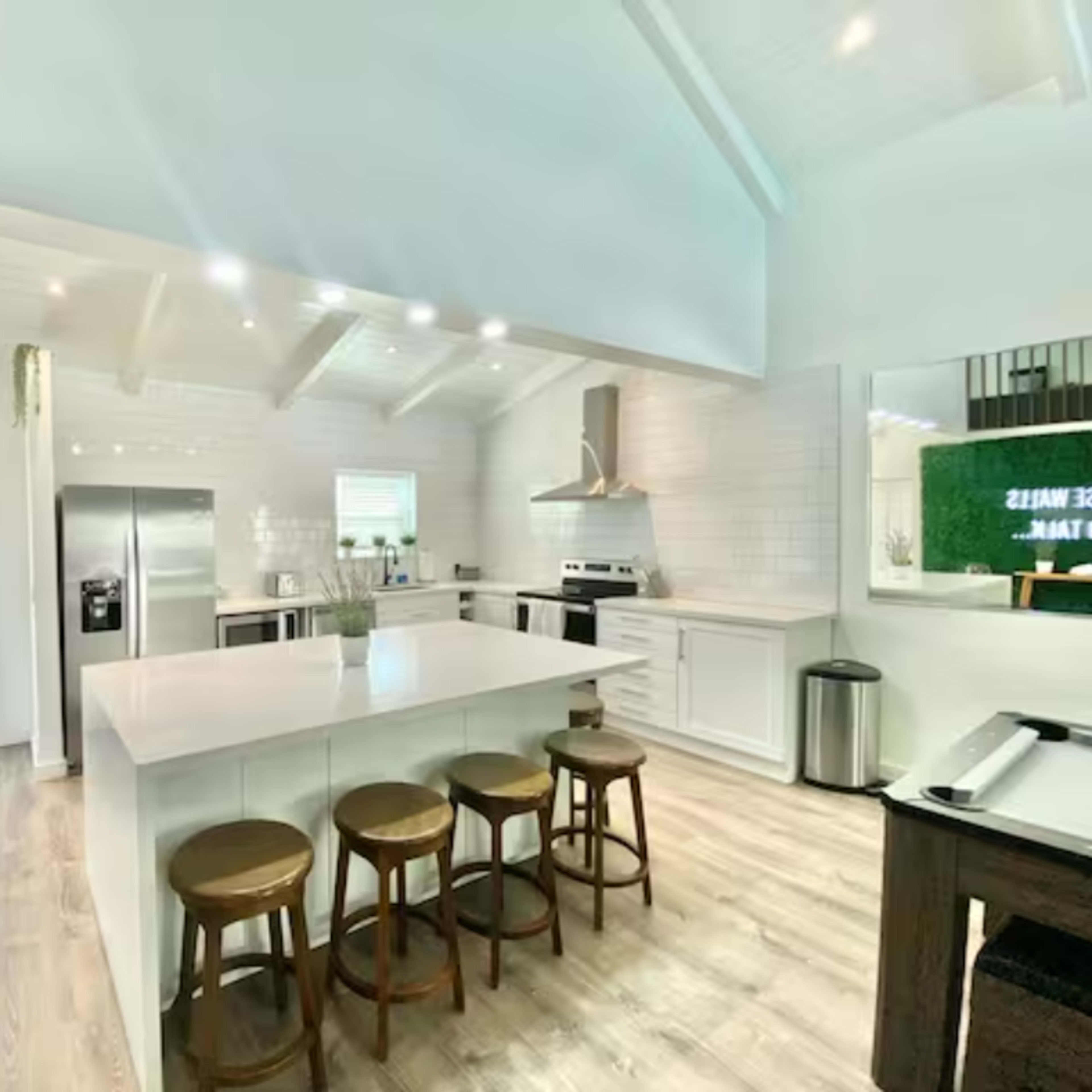A modern kitchen features a large island with wooden stools, stainless steel appliances, and white cabinetry under a vaulted ceiling.