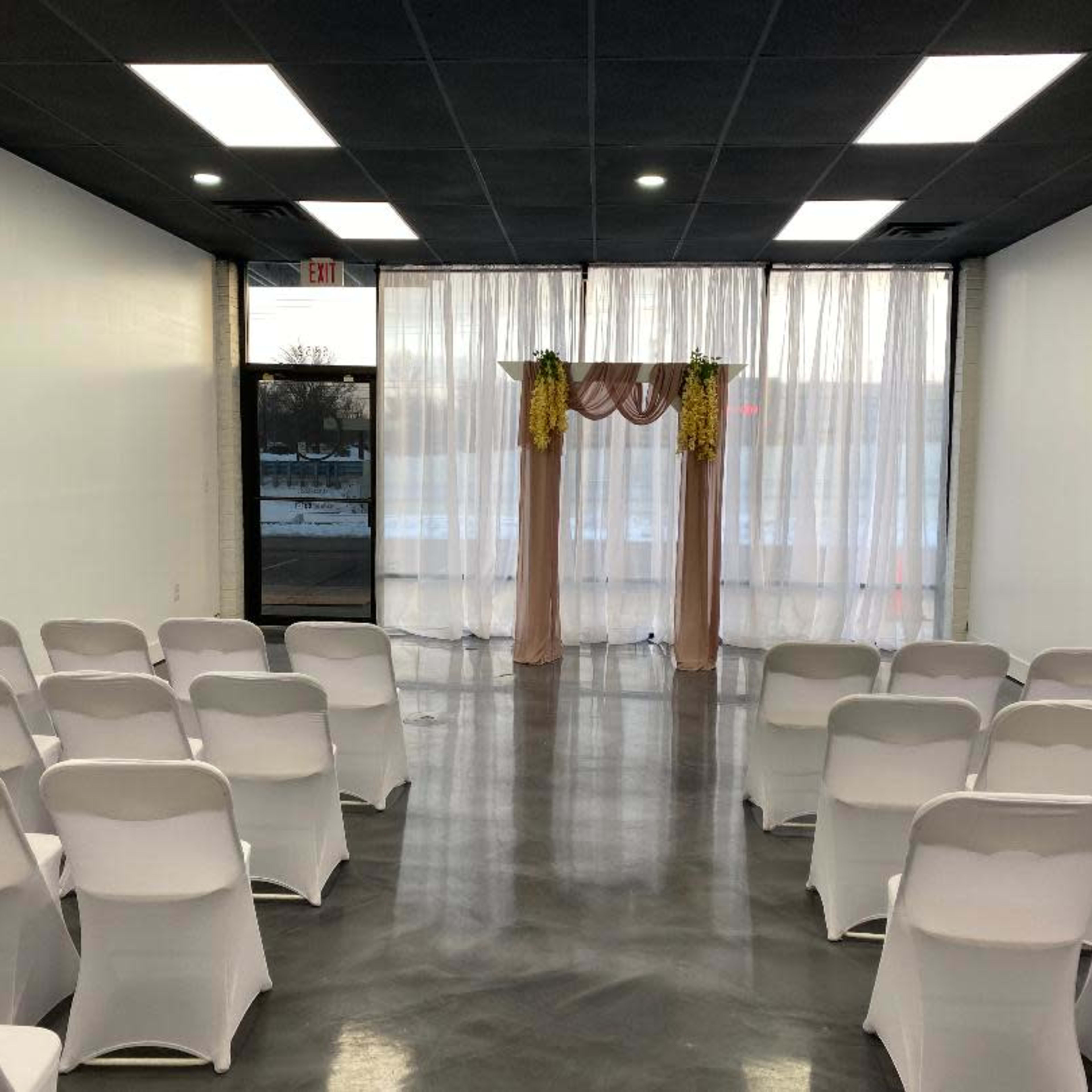 A well-lit event space with rows of white chairs facing a decorative arch draped with fabric at the front, near large windows.