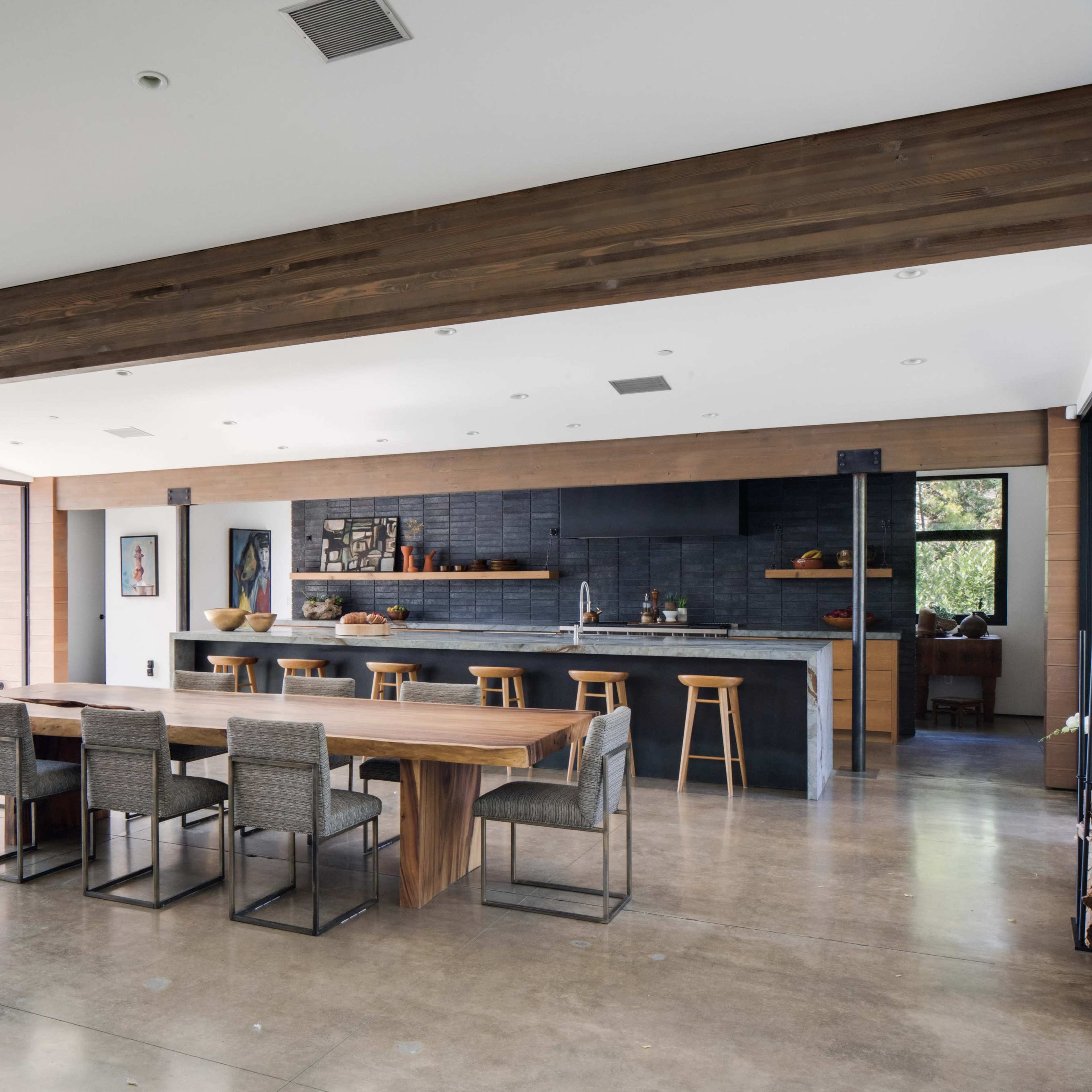A modern open-concept living space featuring a dining area with a long wooden table, a kitchen with dark cabinetry, and large windows overlooking a landscaped exterior.