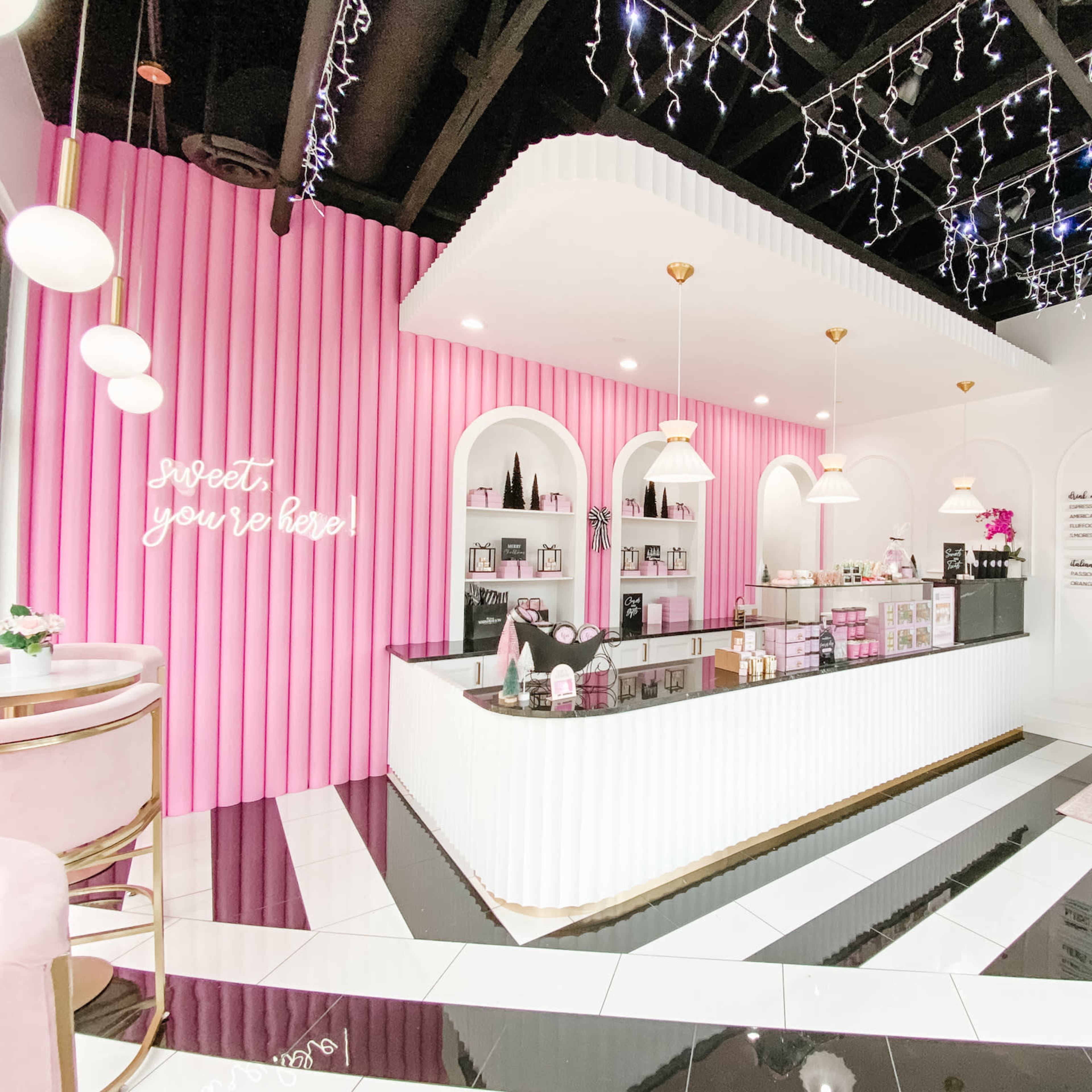 A brightly lit beauty store features a sleek white and black counter, a pink accent wall with shelves, and decorative lighting overhead.