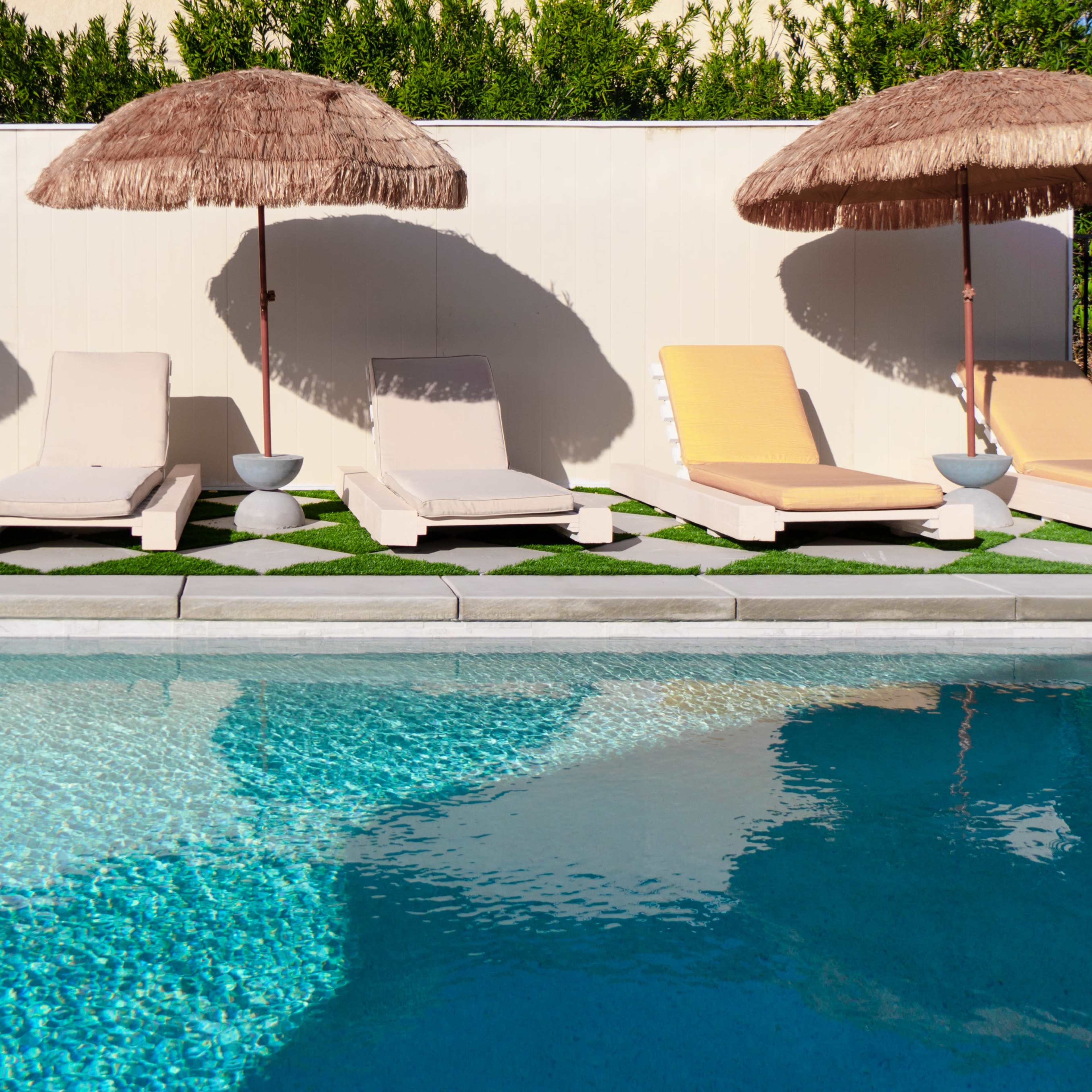 Lounge chairs with umbrellas are arranged beside a swimming pool surrounded by neatly maintained grass and a white wall.
