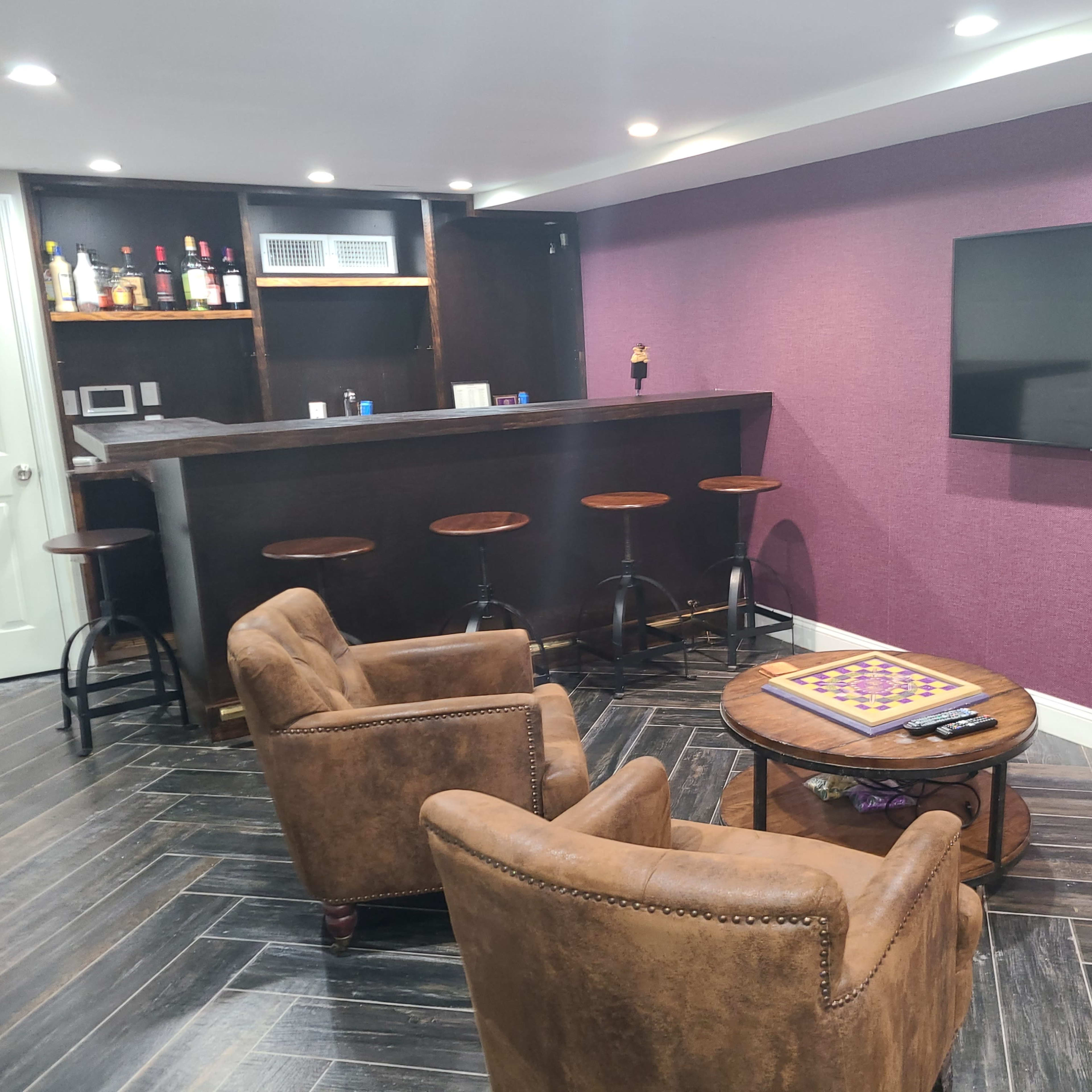 A cozy basement lounge features a bar with high stools, two brown armchairs, a round table with a game board, and a large flat-screen TV mounted on a purple wall.