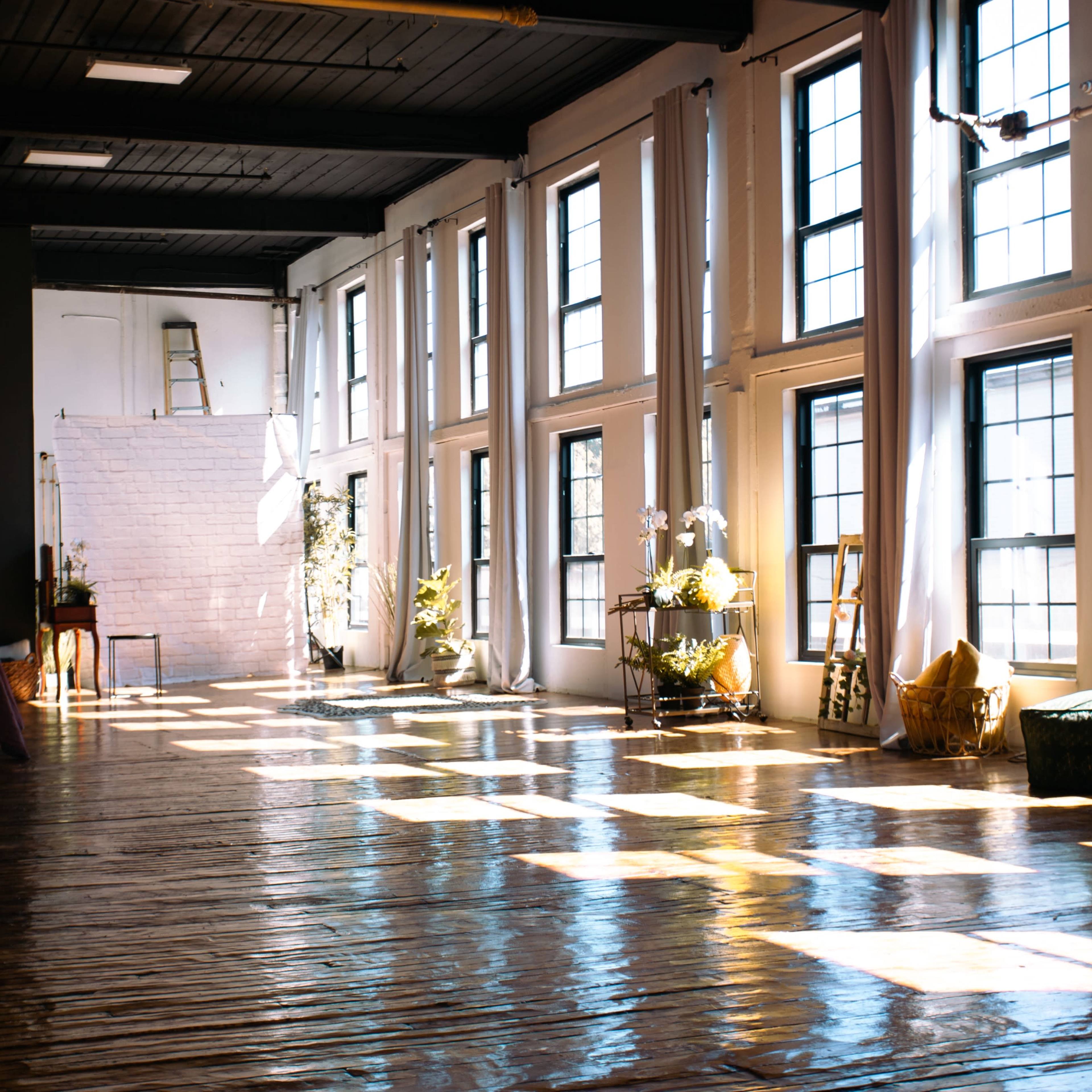 A spacious room with large windows casting shadows on a polished wooden floor, featuring plants and a few pieces of furniture.