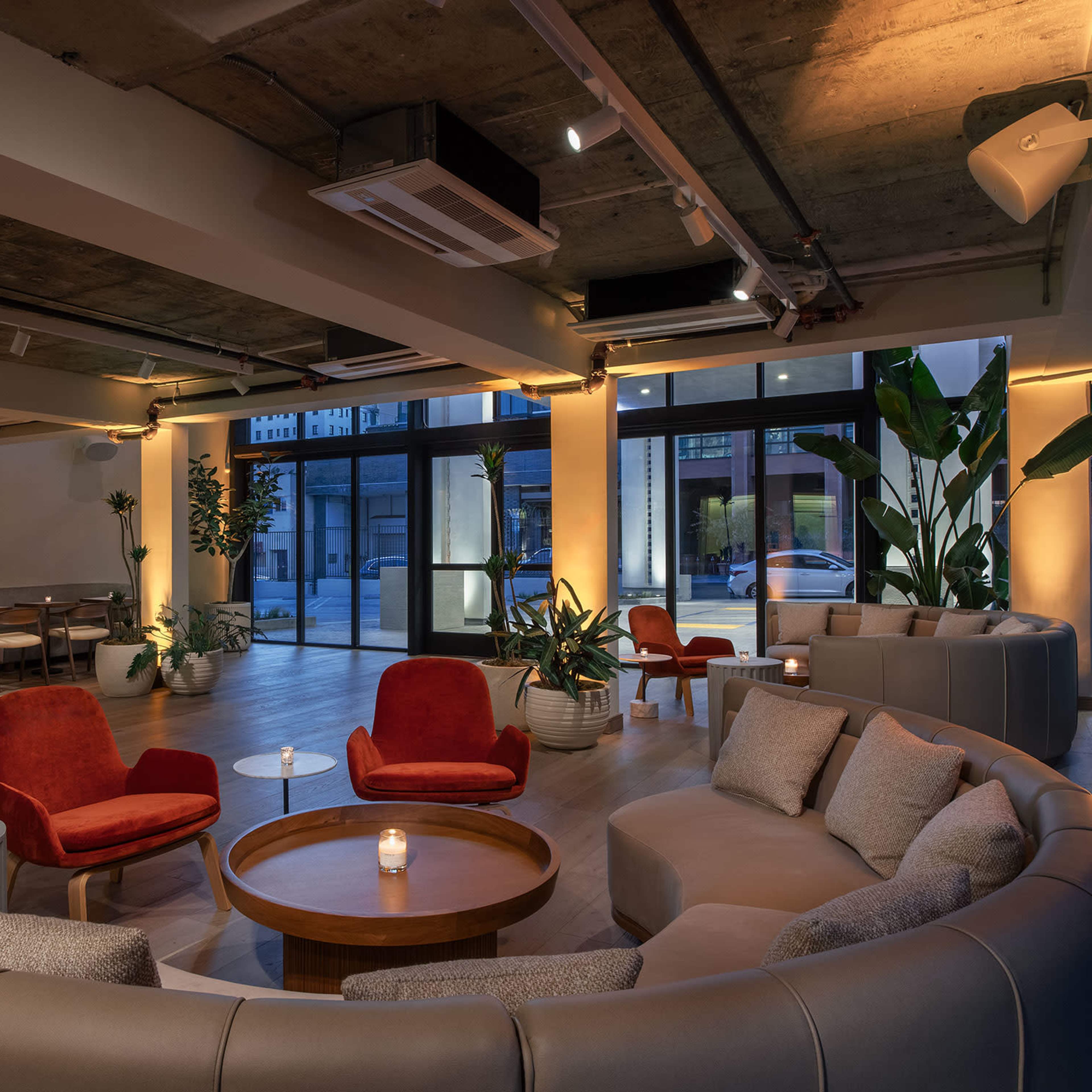 A modern lounge with circular seating, a wooden coffee table, and potted plants, illuminated by warm lighting.