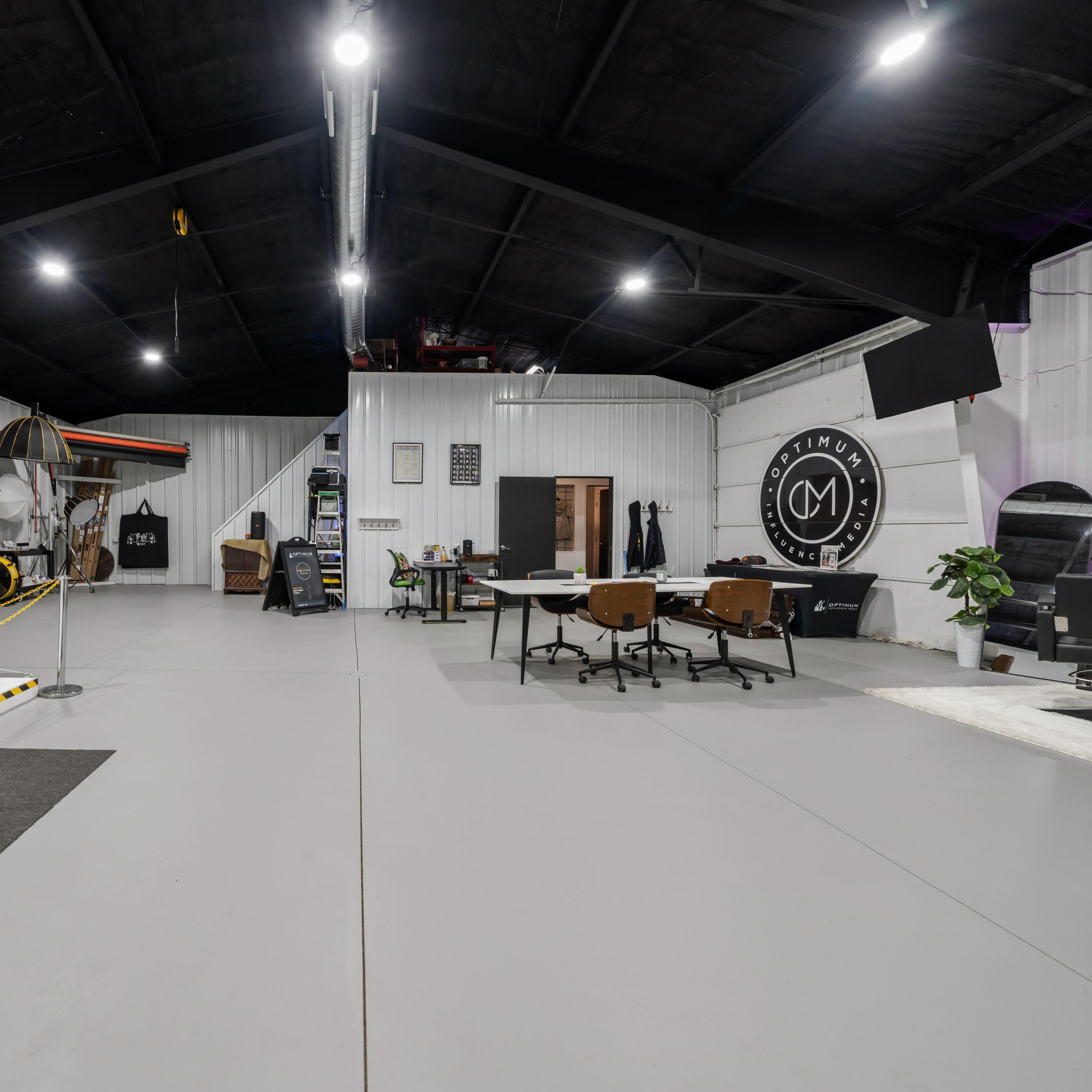 A spacious interior of a modern studio or workspace, featuring a large open area with desks, chairs, and various equipment against a backdrop of white walls and a black ceiling.