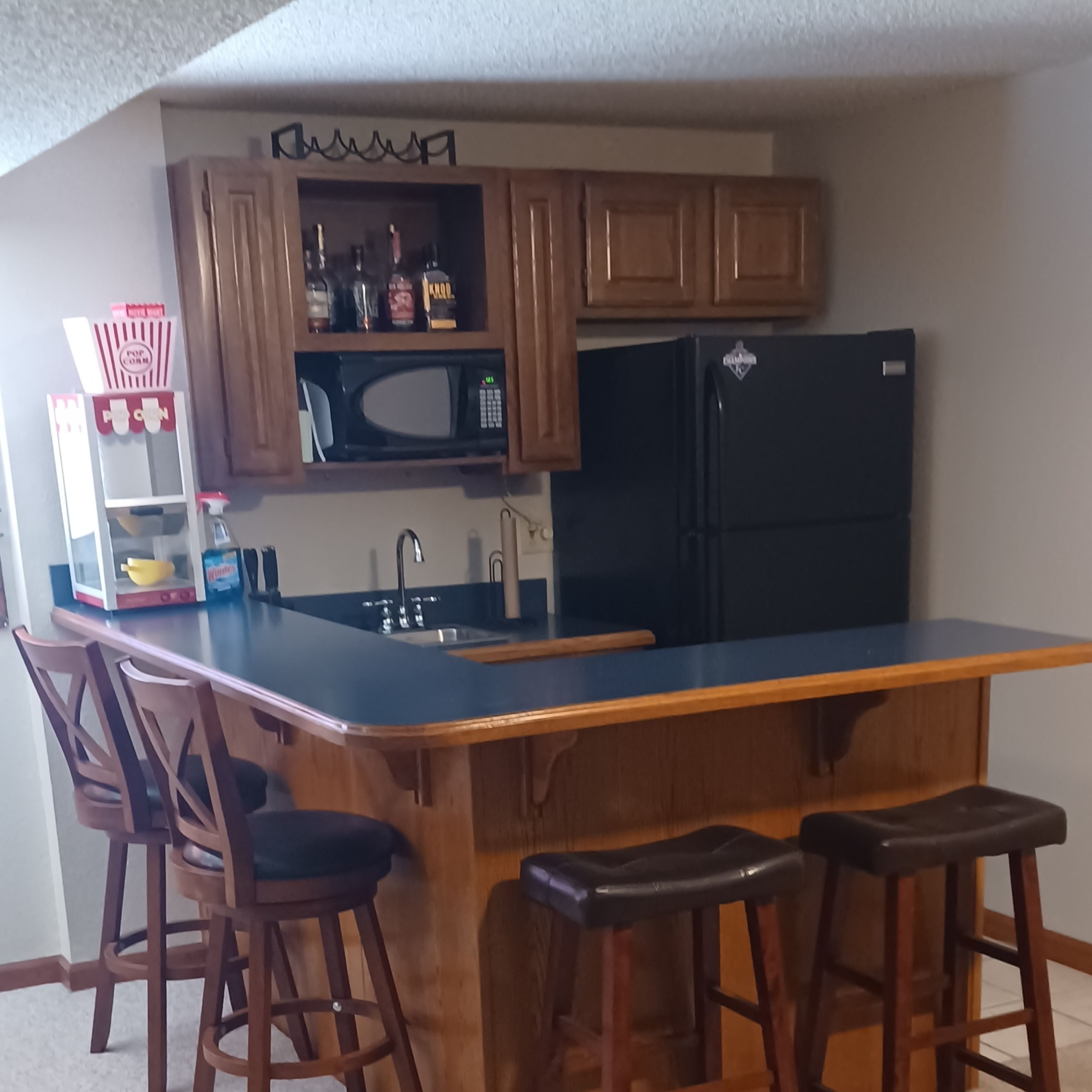 A basement bar area features a wooden counter with five stools, a sink, a refrigerator, and shelves above for drinks.