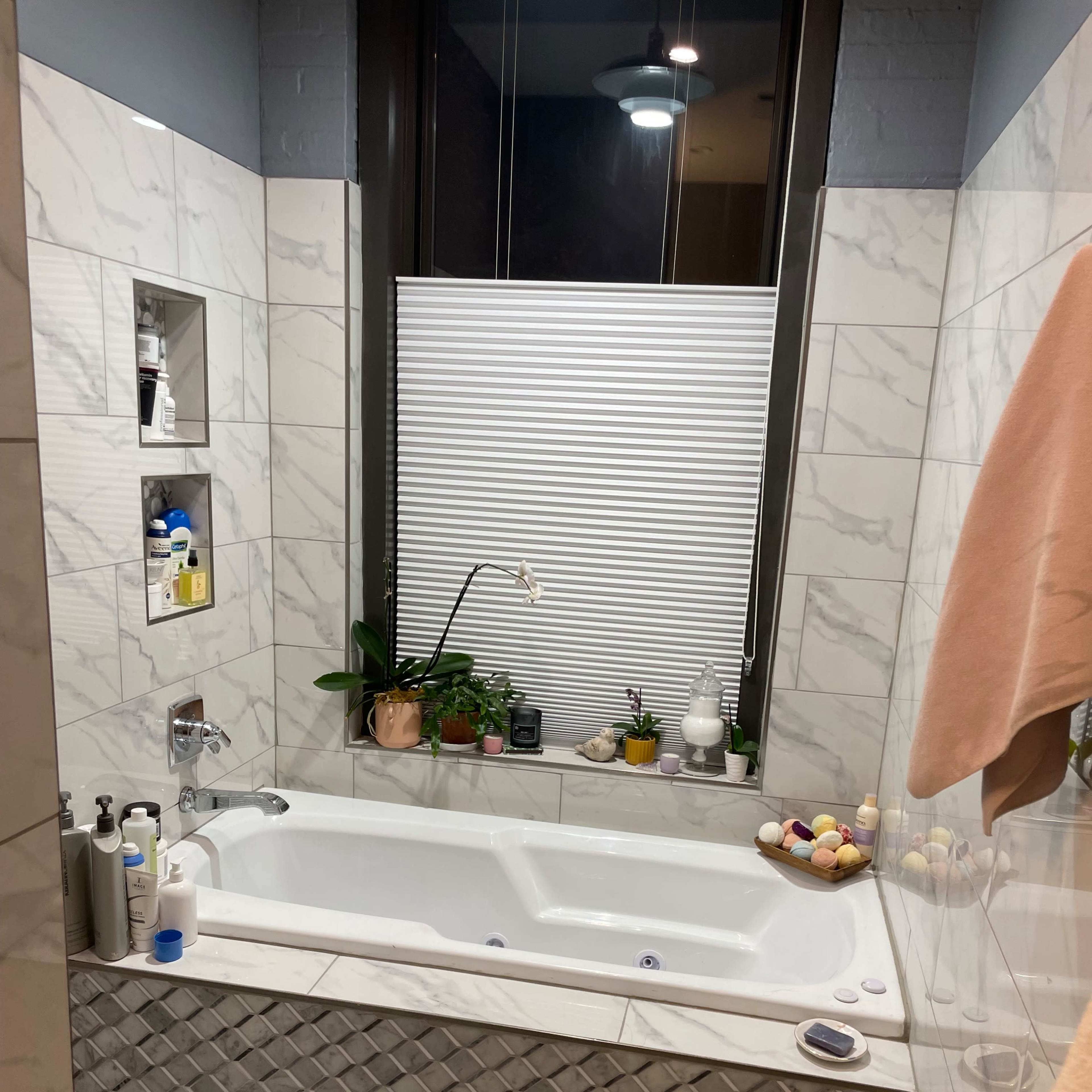 A modern bathroom featuring a large soaking tub surrounded by decorative tiles, a window with blinds, and several potted plants on the windowsill.
