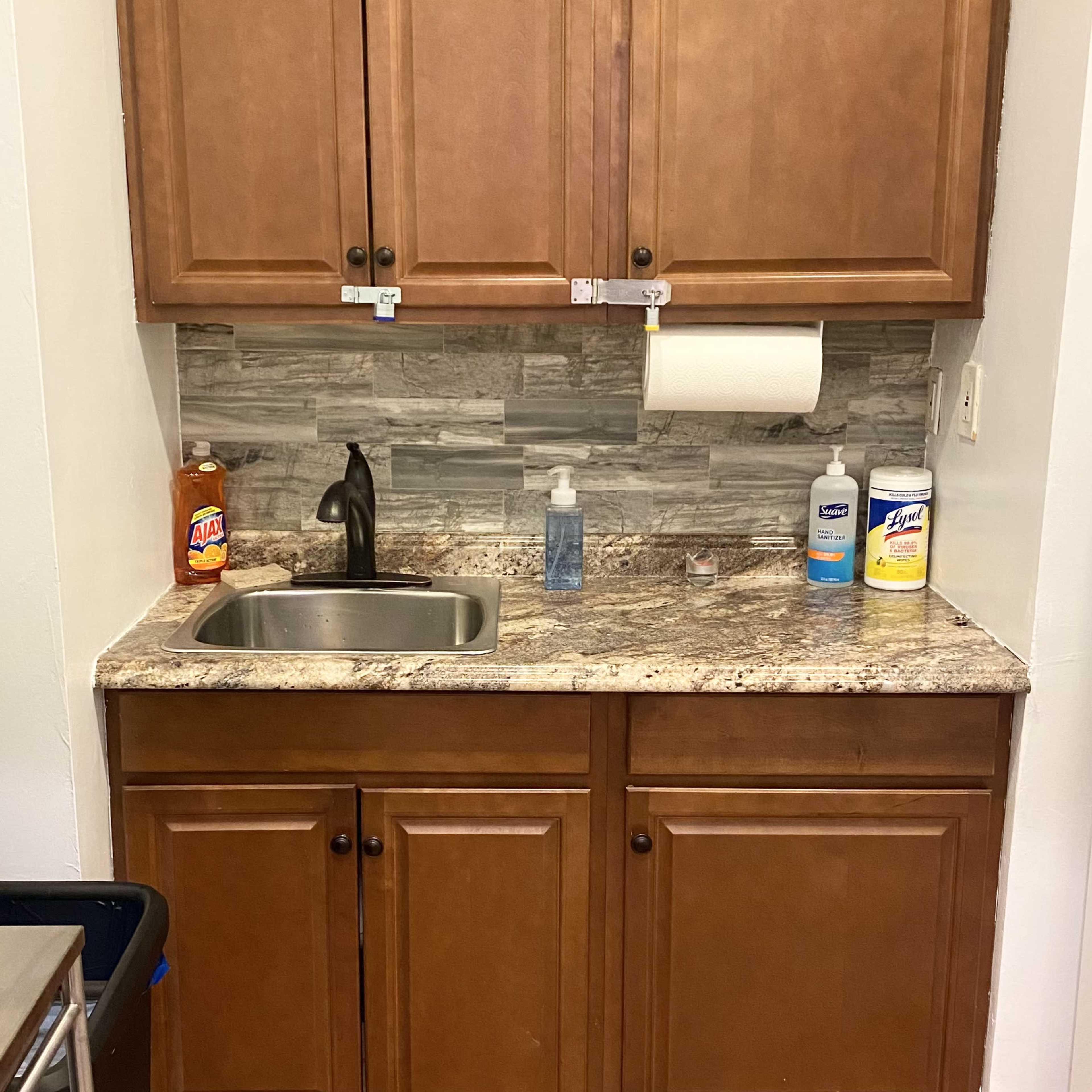 A small kitchen area with a stainless steel sink, a granite countertop, various cleaning supplies, and brown wooden cabinets above.