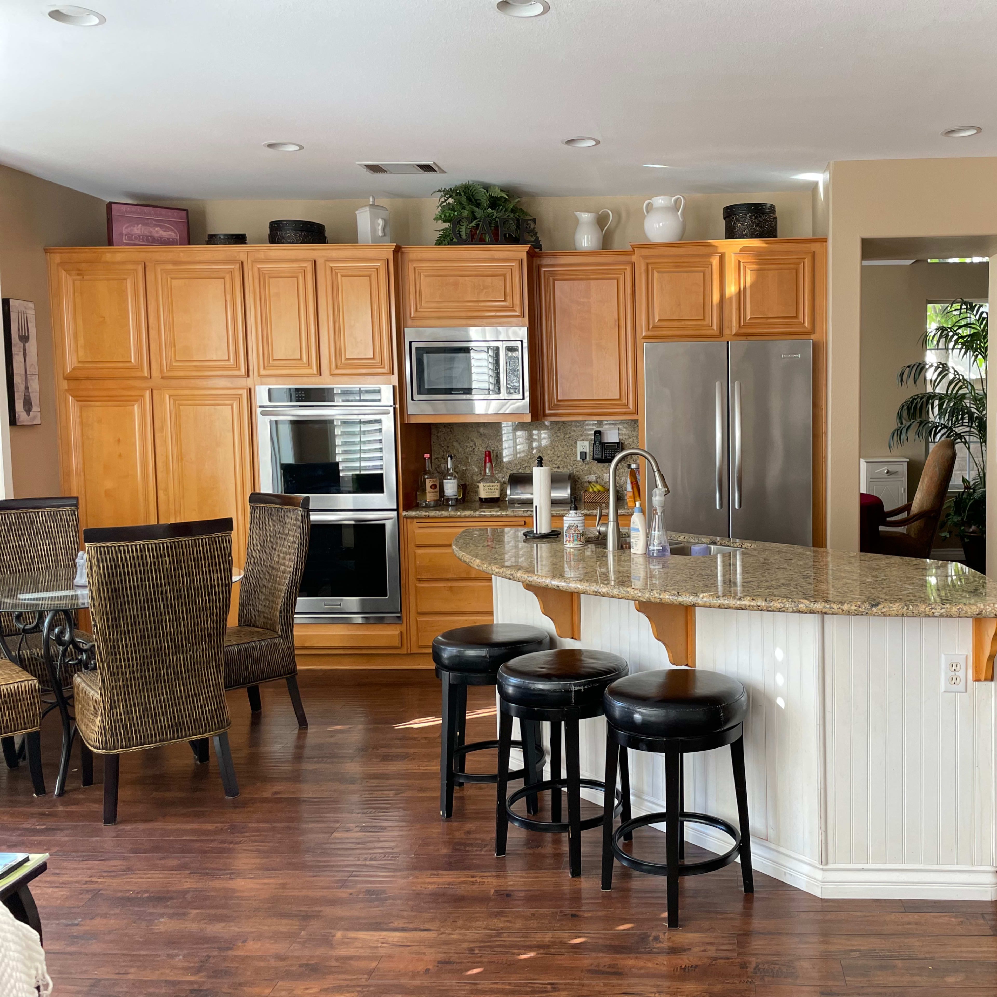 A kitchen with wooden cabinets, a granite countertop island, black bar stools, and a dining area with wicker chairs.