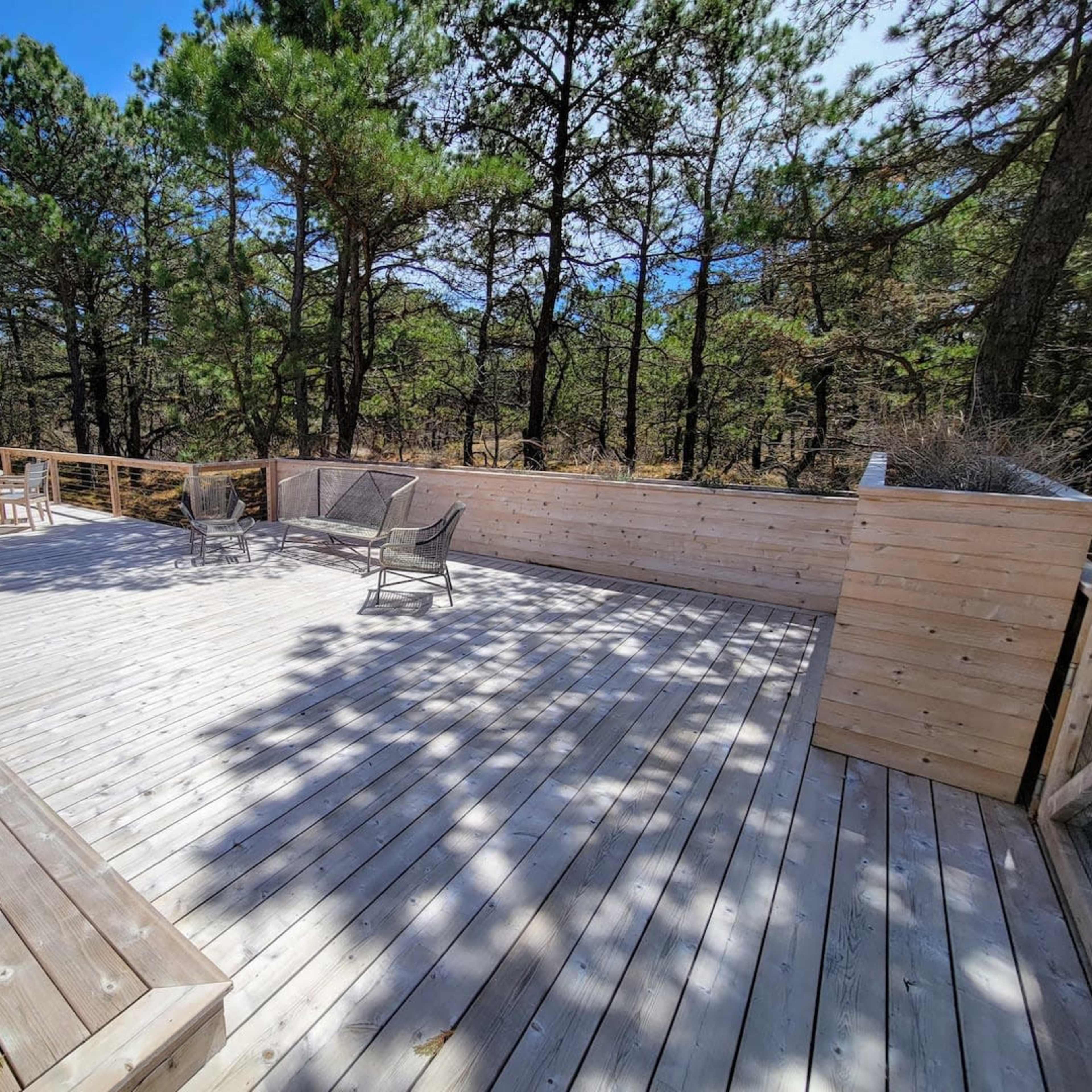 A wooden deck surrounded by trees features a few chairs and tables, creating an outdoor seating area.