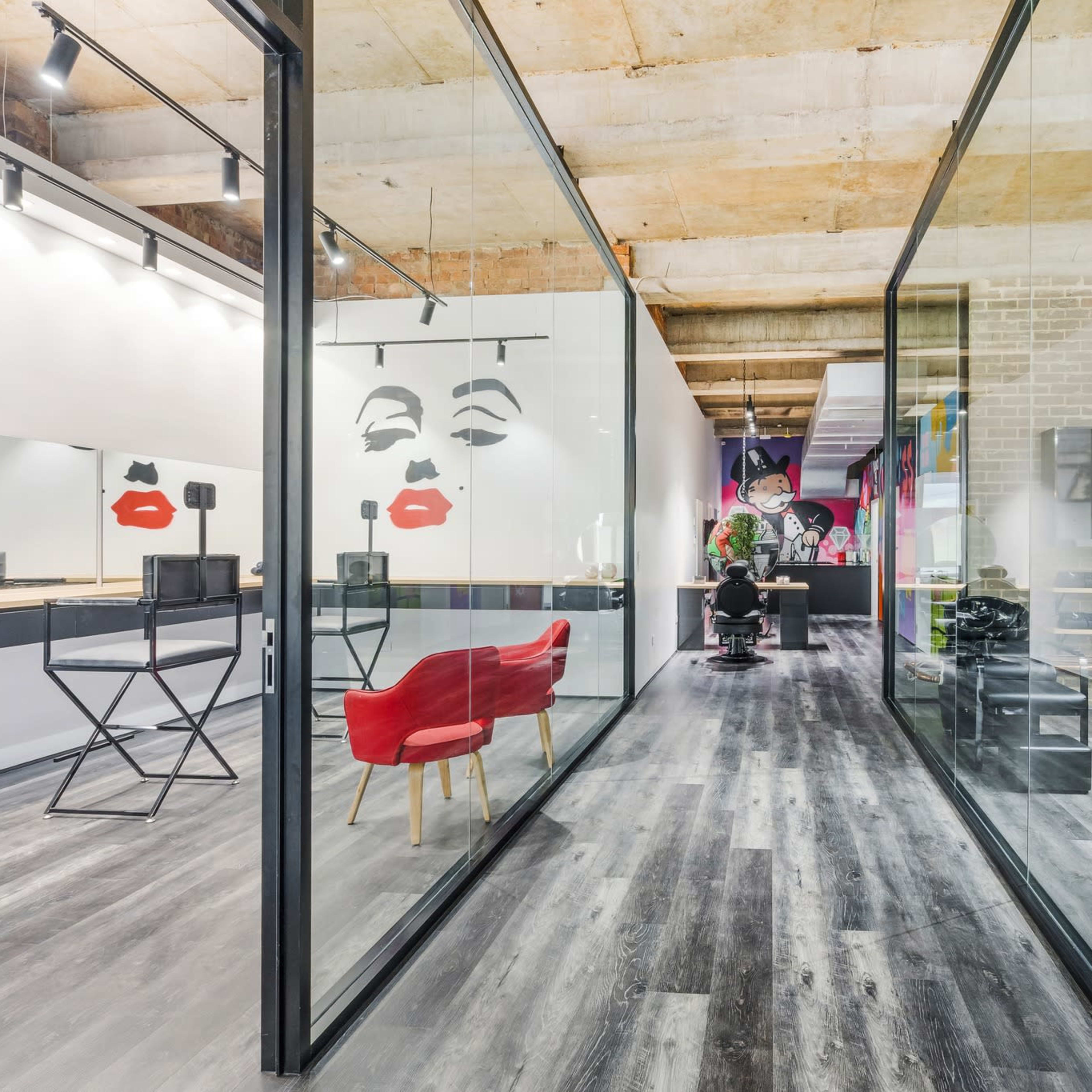 A modern salon interior with glass partitions, featuring colorful wall art and an area with styling stations and furniture.