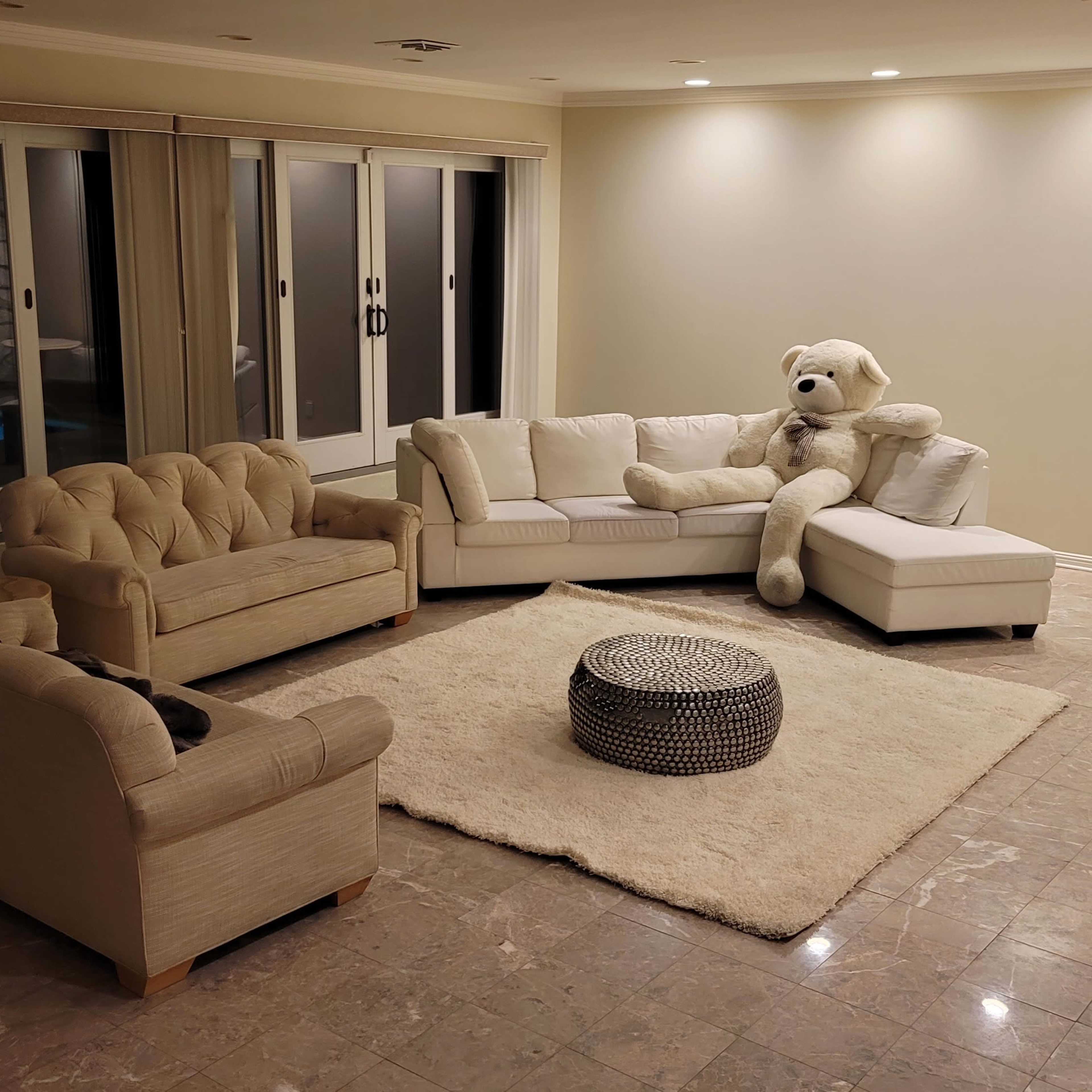 A spacious living room features a large white sectional sofa, two beige tufted sofas, a round coffee table, a plush area rug, and a giant teddy bear sitting on the sectional near large glass doors.