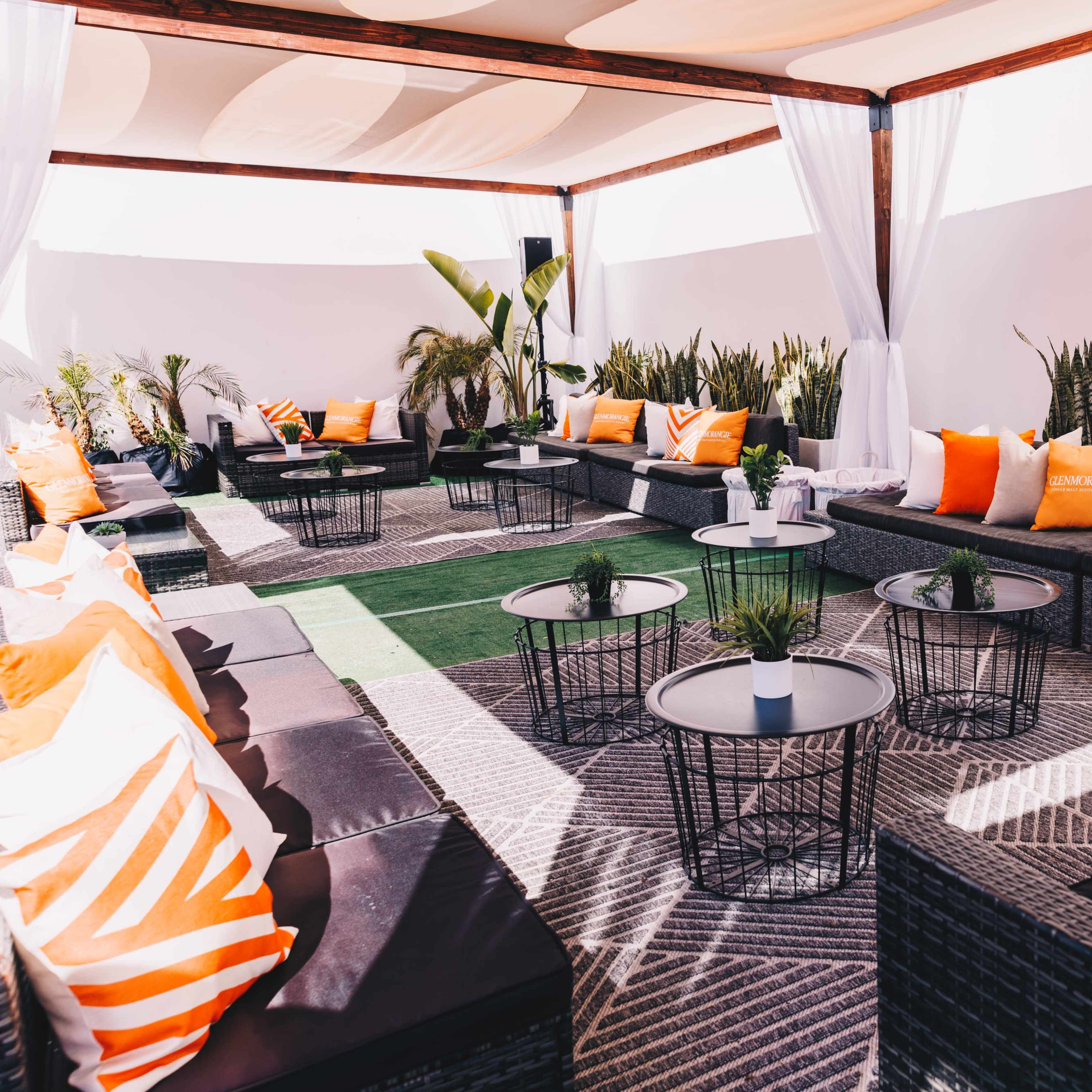 A modern outdoor lounge area with black wicker furniture, orange and white cushions, and small tables, surrounded by plants.