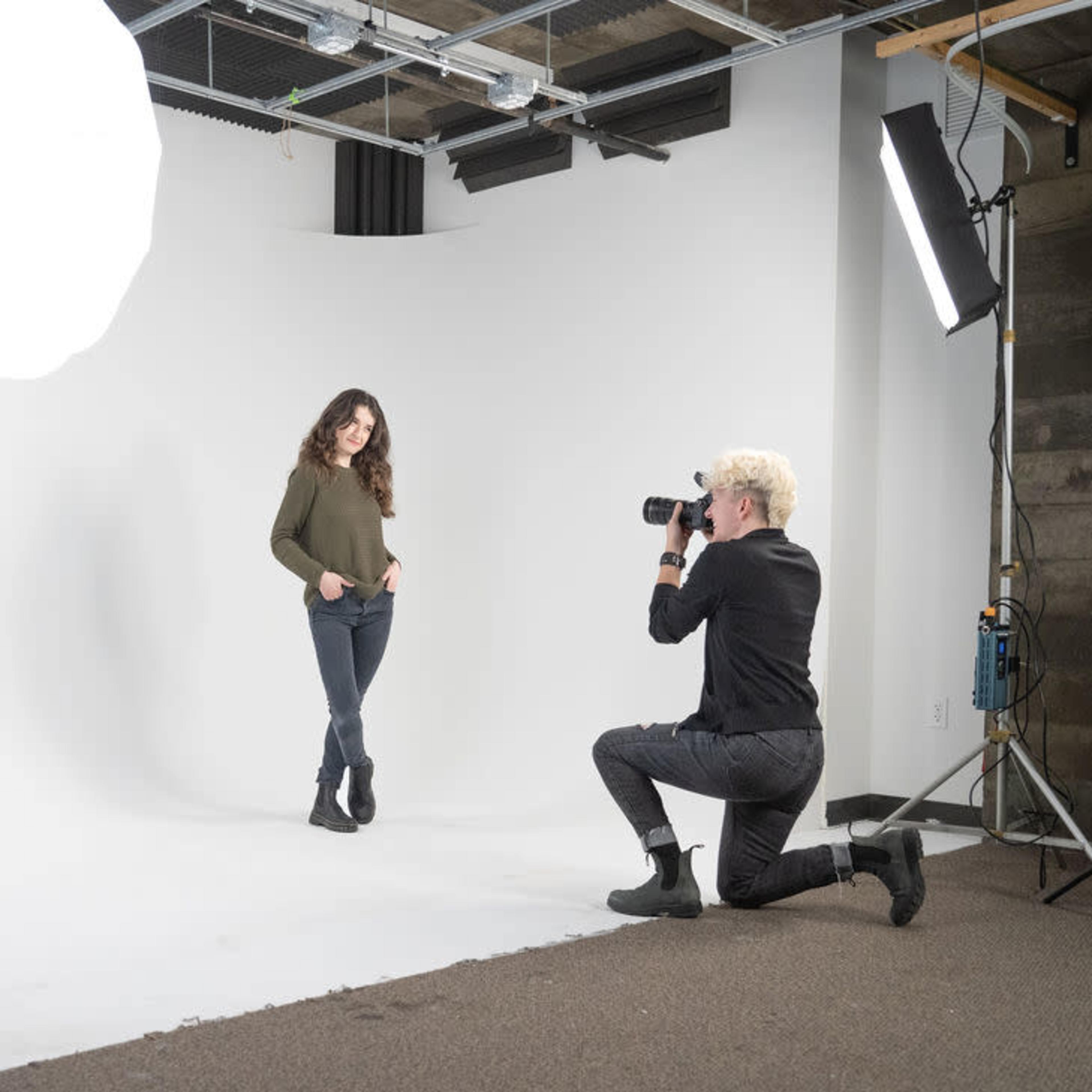 A photographer kneels while taking pictures of a model in a simple studio setup with a white backdrop and lighting equipment.