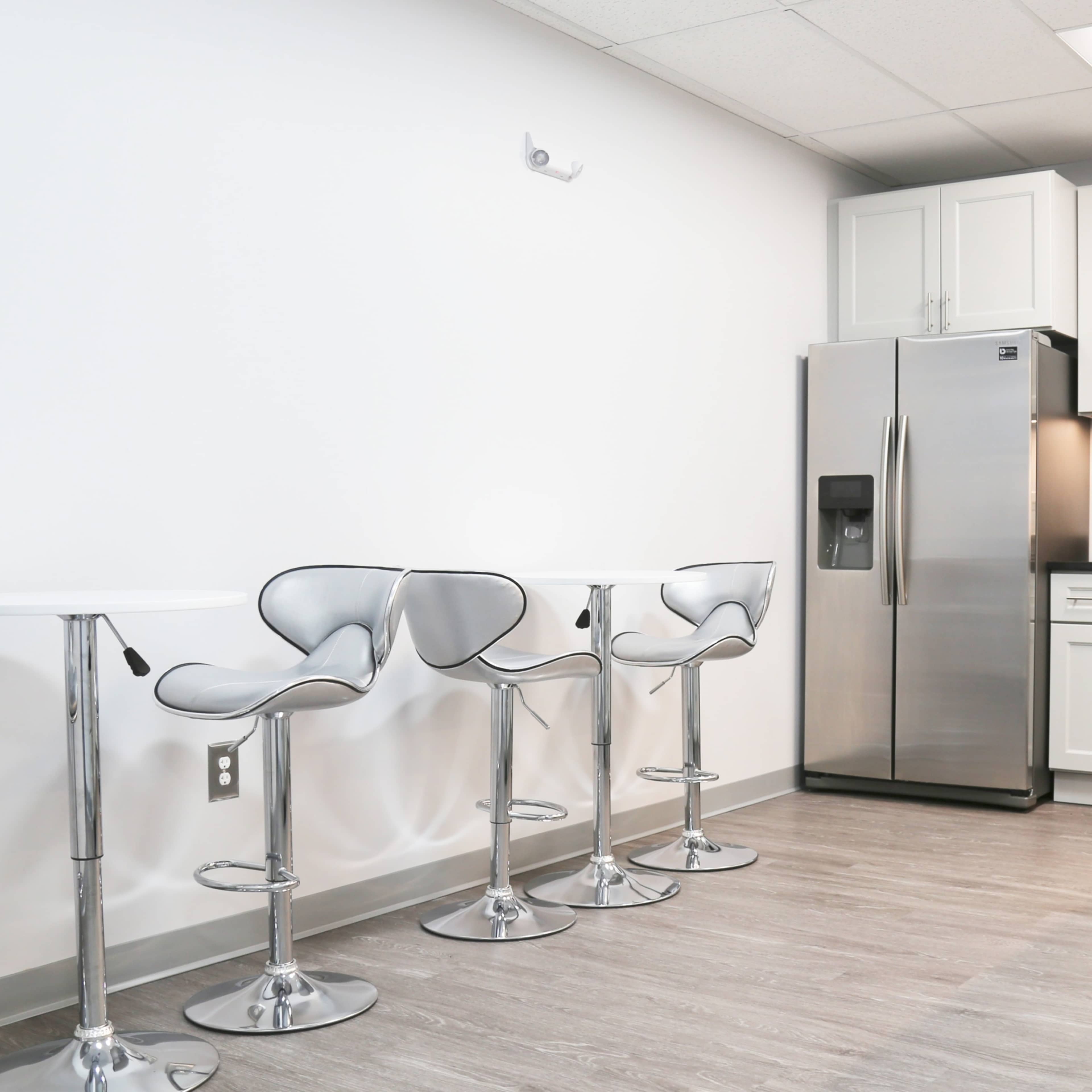 A modern kitchen area with a stainless steel refrigerator, a microwave, and four high-backed chrome stools arranged around two sleek white tables.