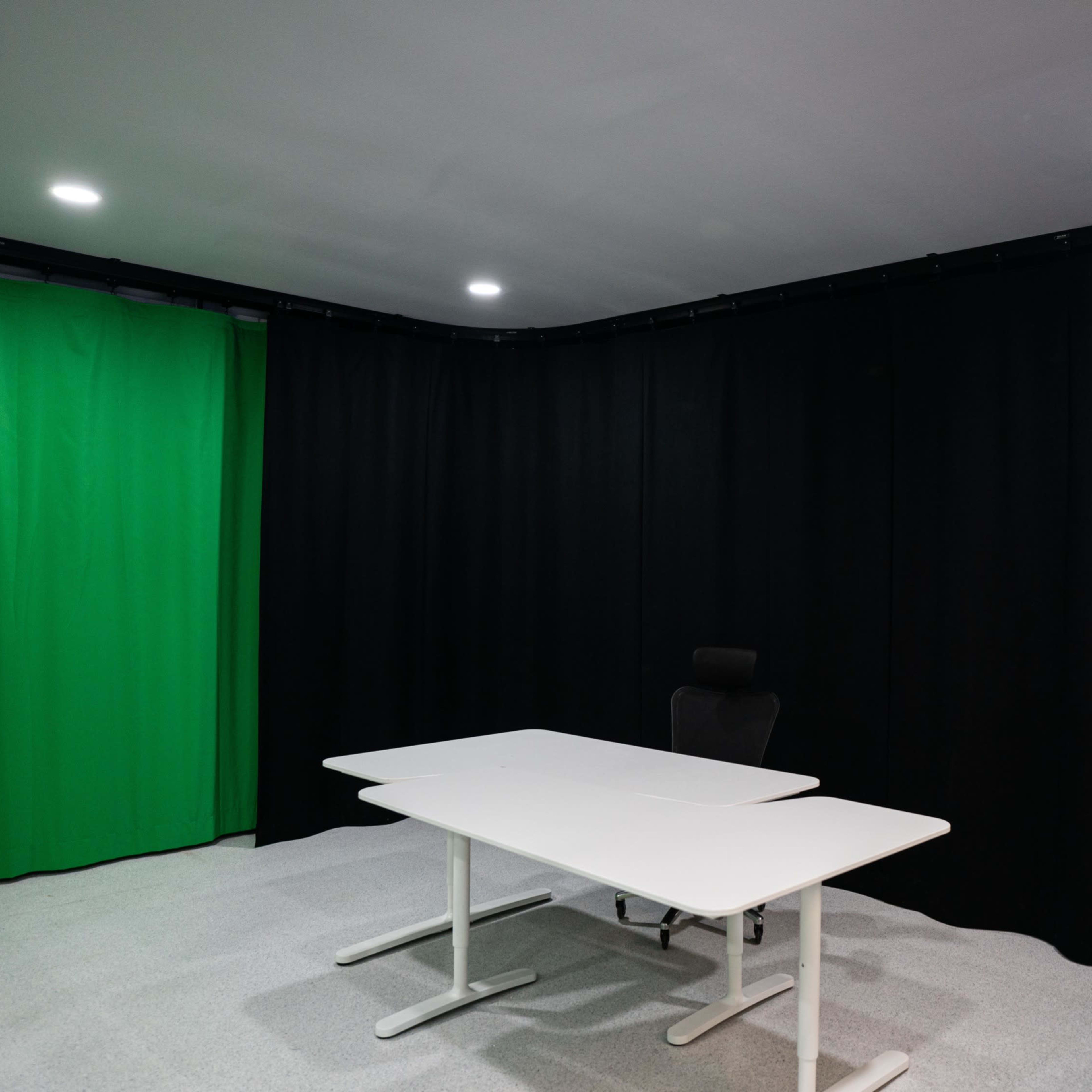 A small studio space with a green screen, black curtains, and a white table with a chair.