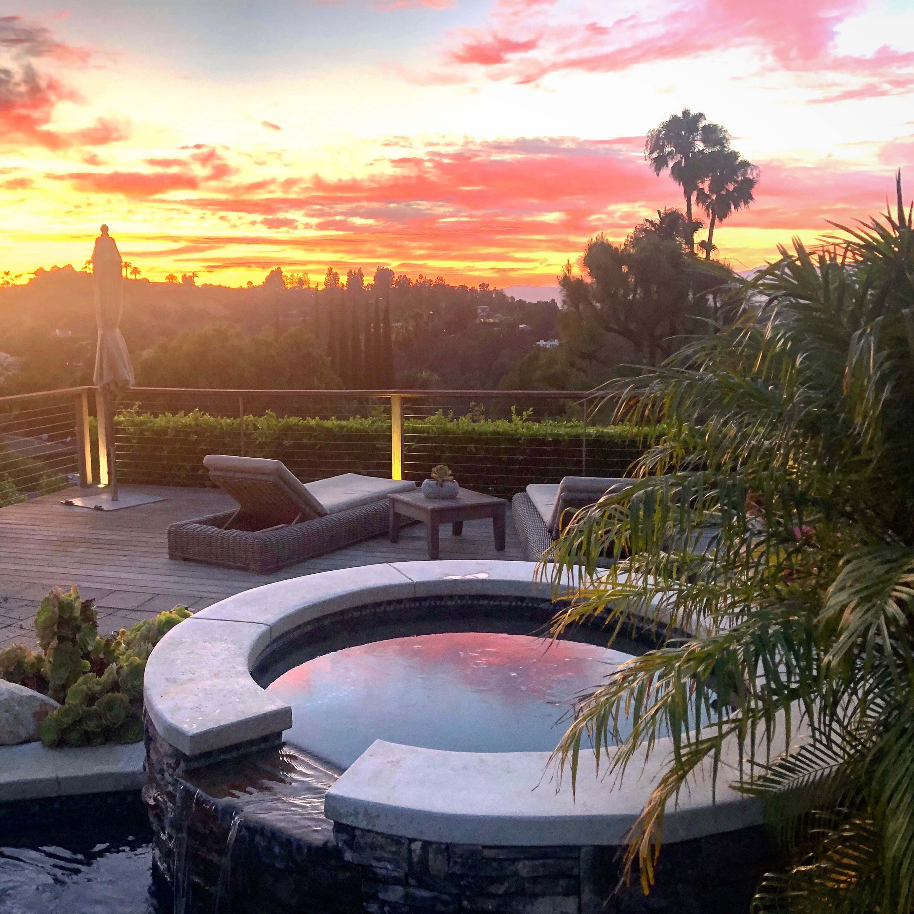 A hot tub surrounded by greenery and patio furniture, with a vibrant sunset in the background.