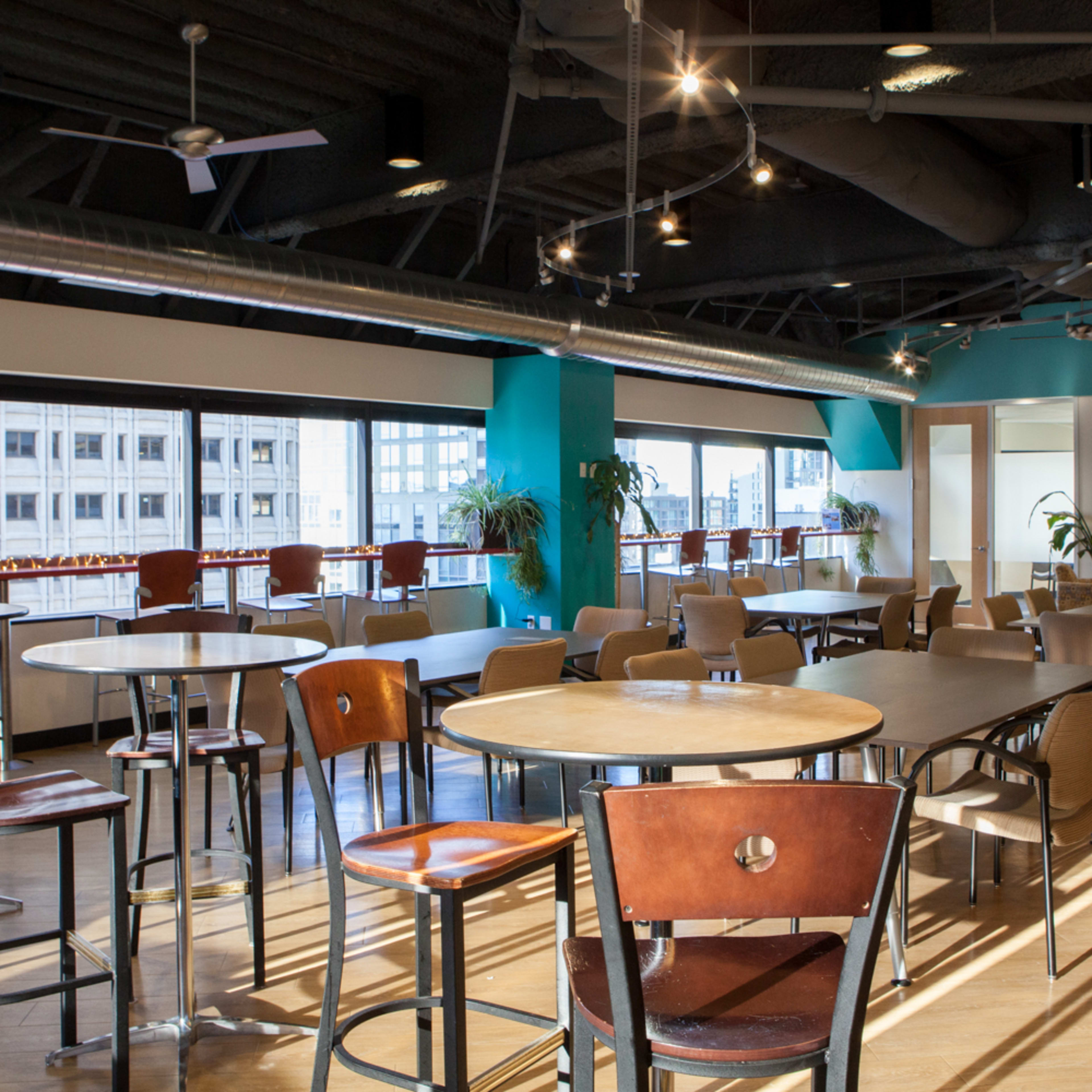 A spacious, modern lounge area with multiple round and square tables, surrounded by large windows and potted plants.