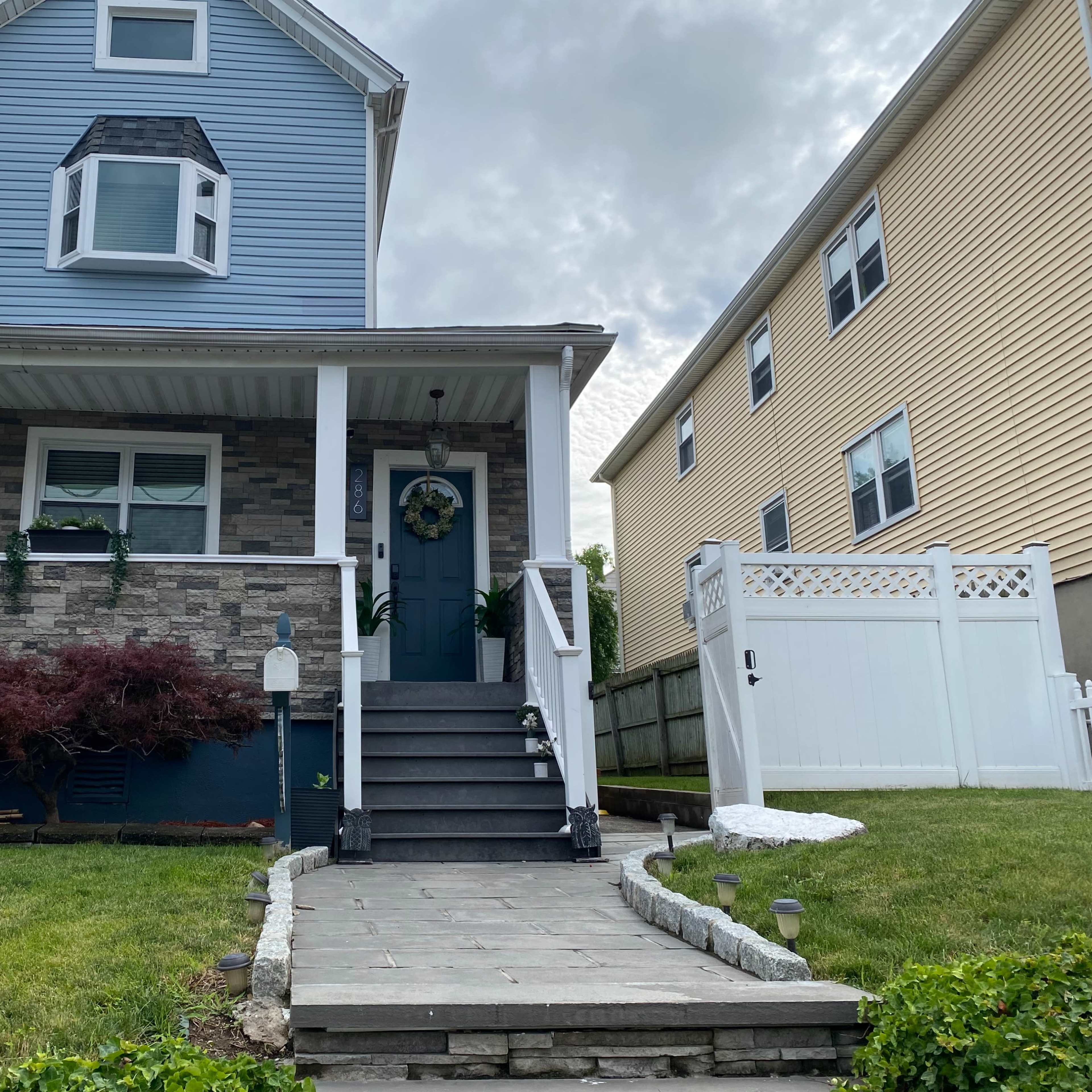 A two-story blue house with stone accents features a front porch, a set of stairs, and a well-maintained lawn, bordered by a white fence.