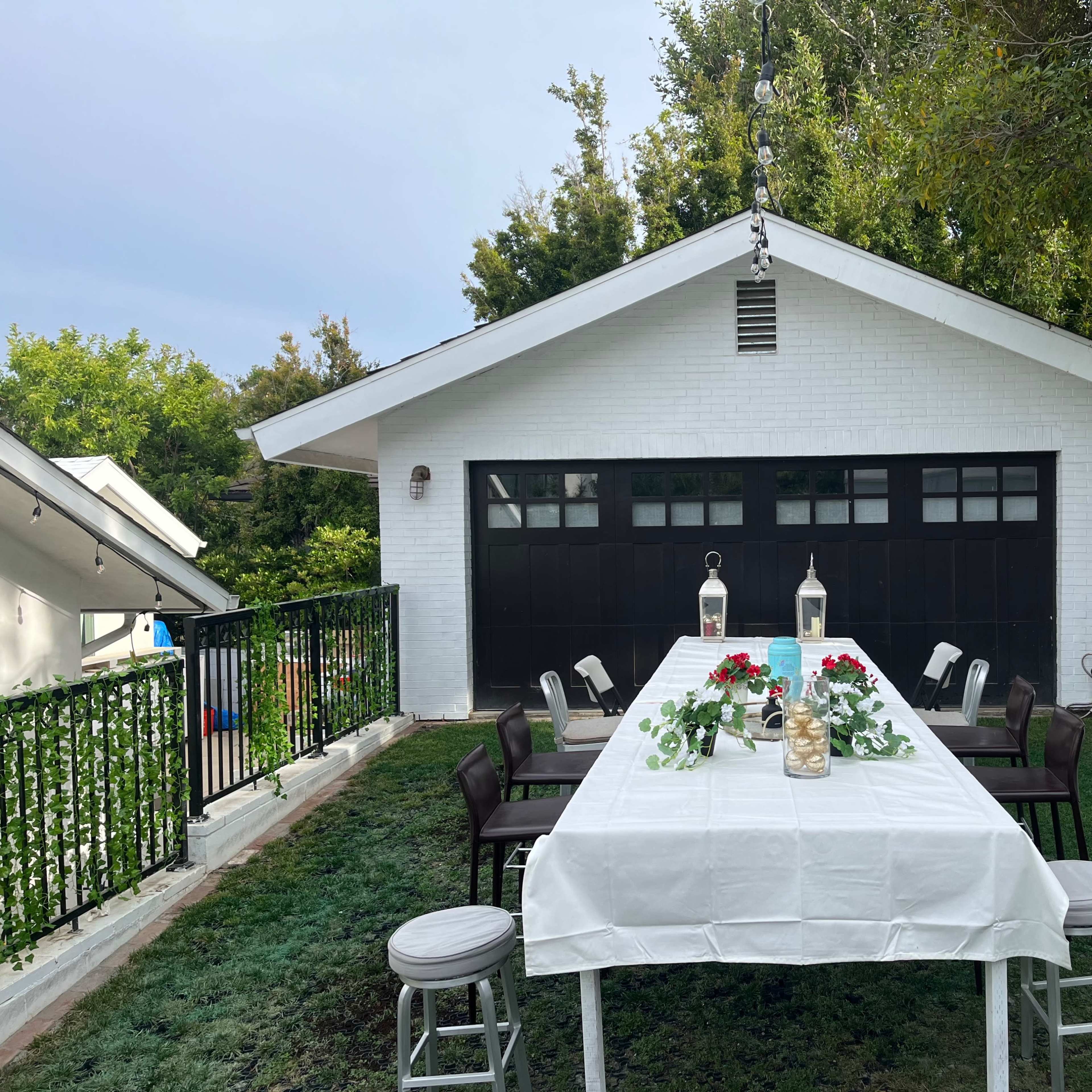 A long table is set outside in front of a white garage, surrounded by green grass and trees.