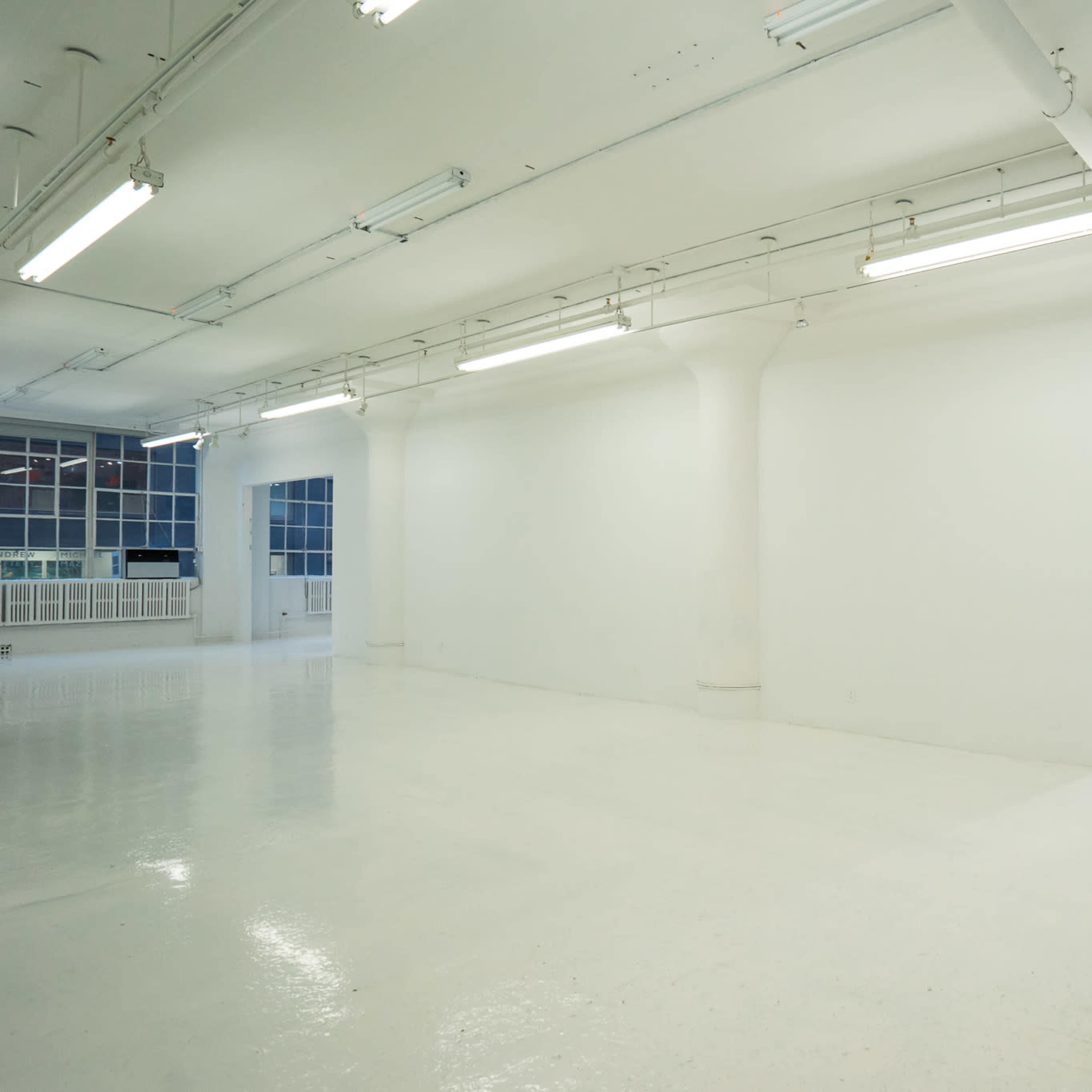A spacious, empty room with white walls and a glossy white floor, illuminated by overhead fluorescent lights.