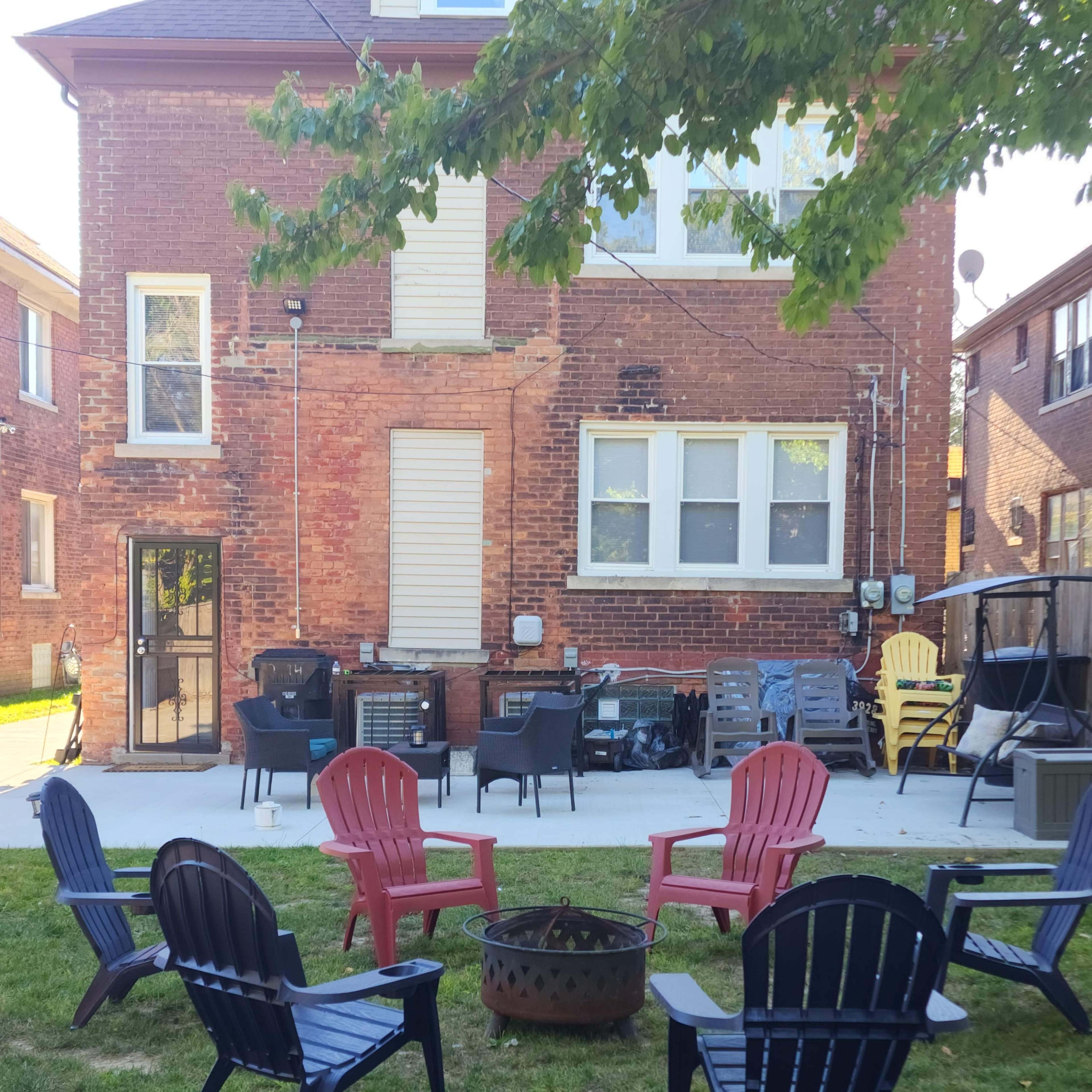 A backyard features a circular arrangement of chairs around a fire pit, with a brick structure and additional seating in the background.