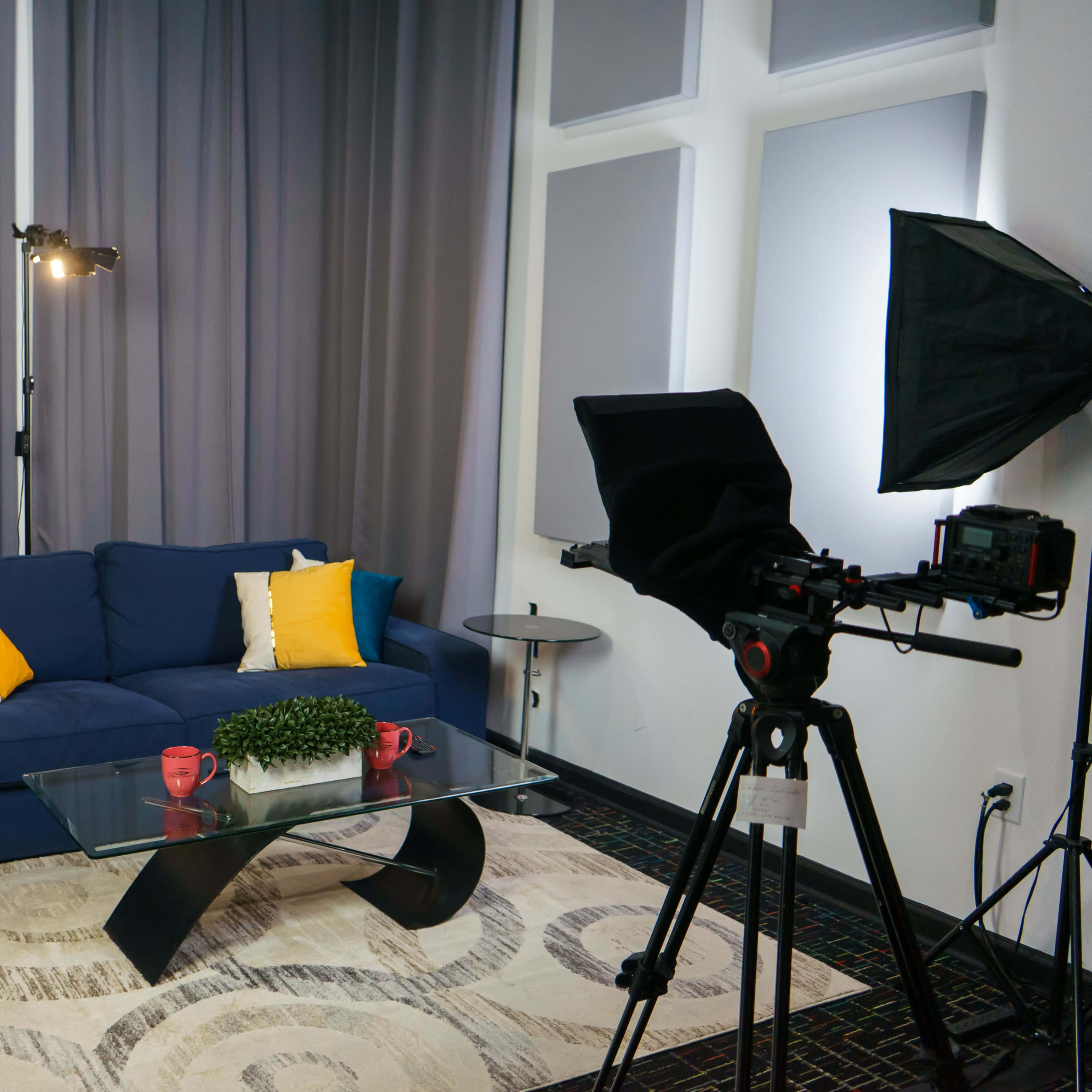 A modern living room setup with a blue sofa, decorative pillows, a glass coffee table, and a camera on a tripod facing the seating area.