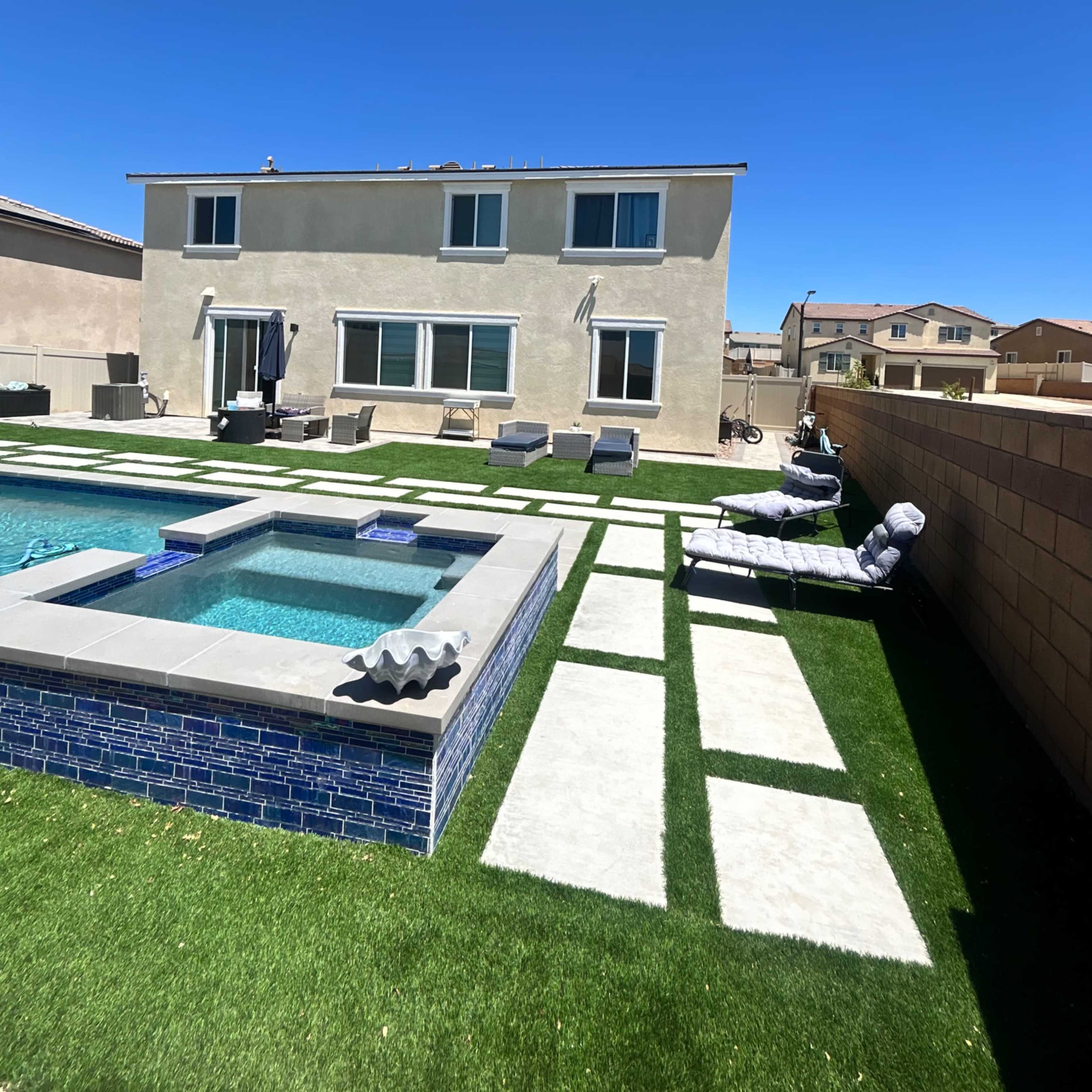 A backyard features a swimming pool with a spa and modern landscaping, including artificial grass and stone pathways, with a two-story house in the background.