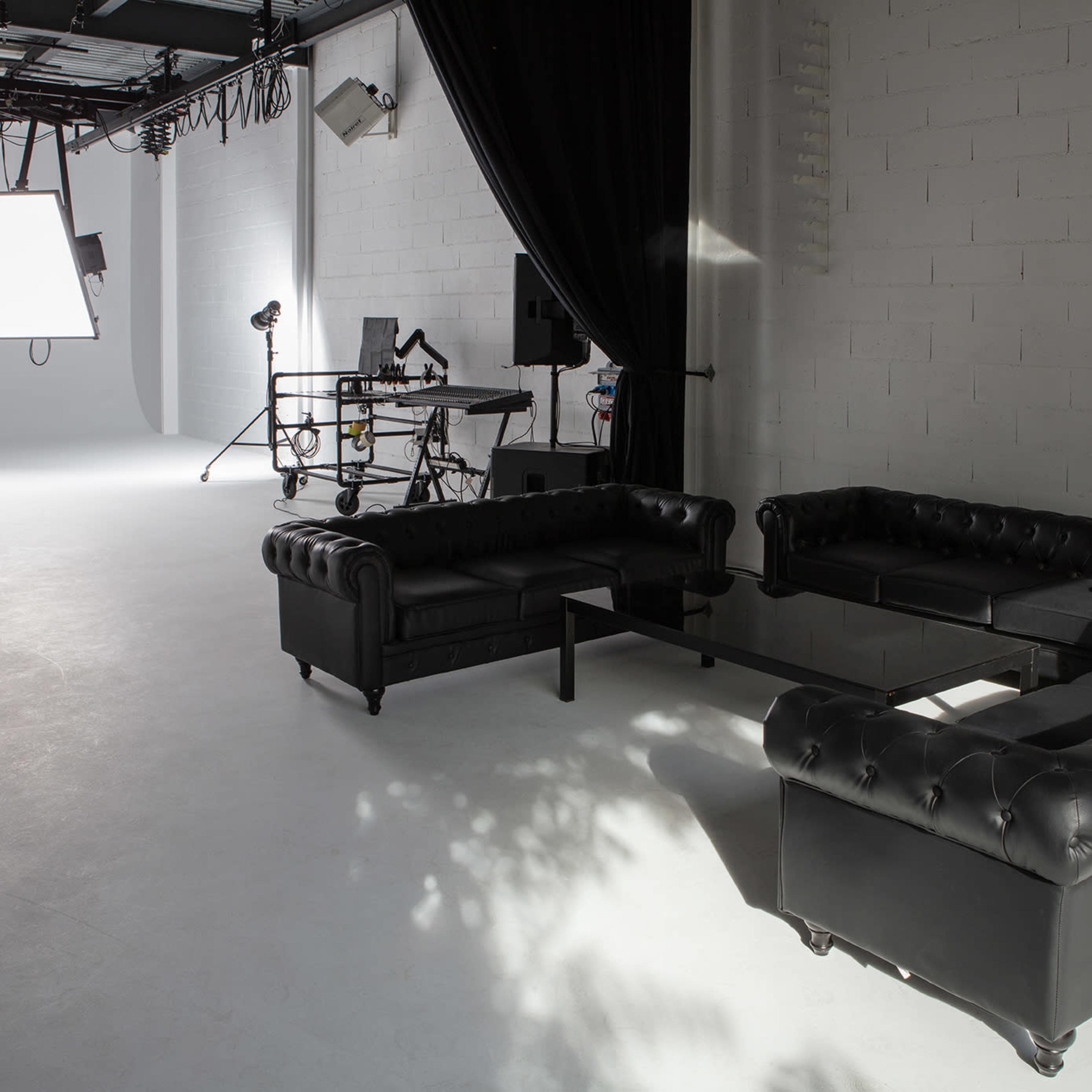 A modern studio setup featuring a black leather sectional couch, a glass coffee table, and various lighting equipment in a minimalist space.