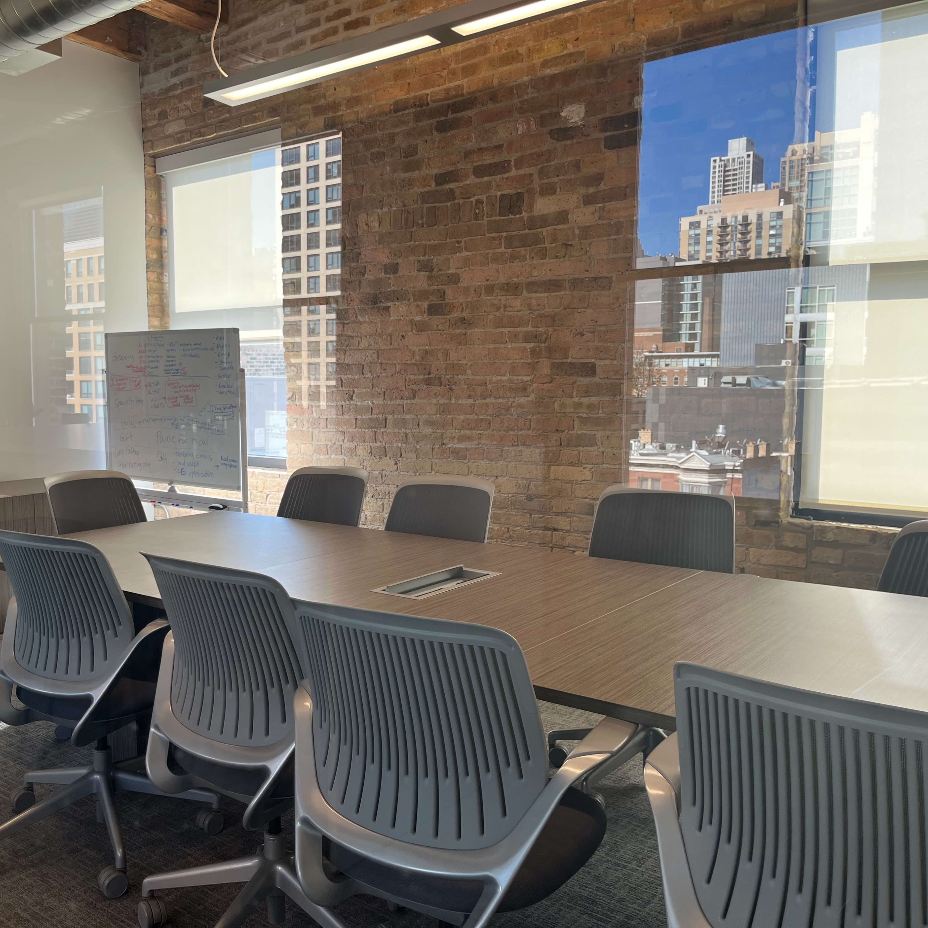 A modern conference room features a large rectangular table surrounded by gray chairs, with a whiteboard and a wall-mounted screen visible in a well-lit space with large windows.