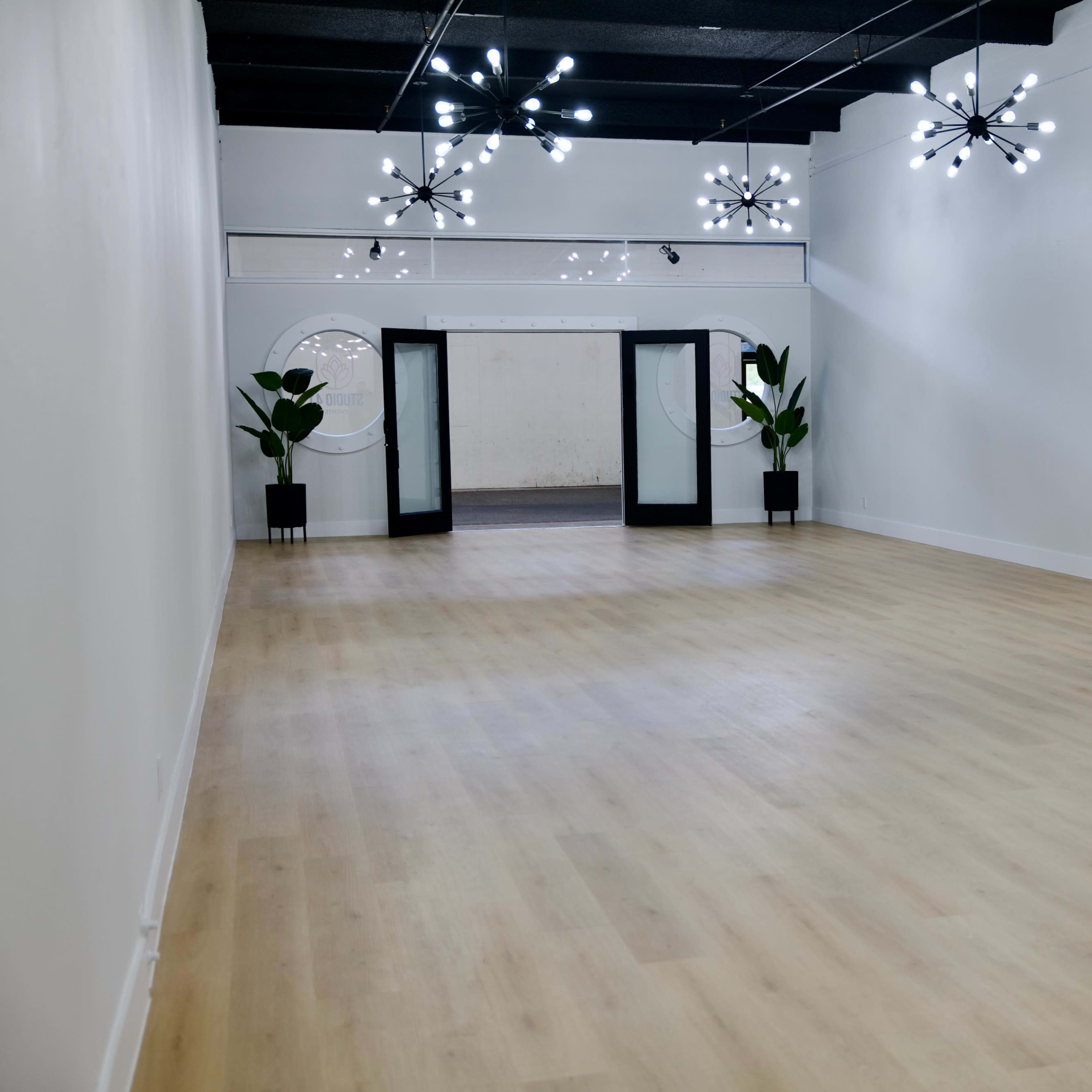 A spacious, empty room with light wooden flooring, featuring modern chandeliers and two large glass doors leading to another area.
