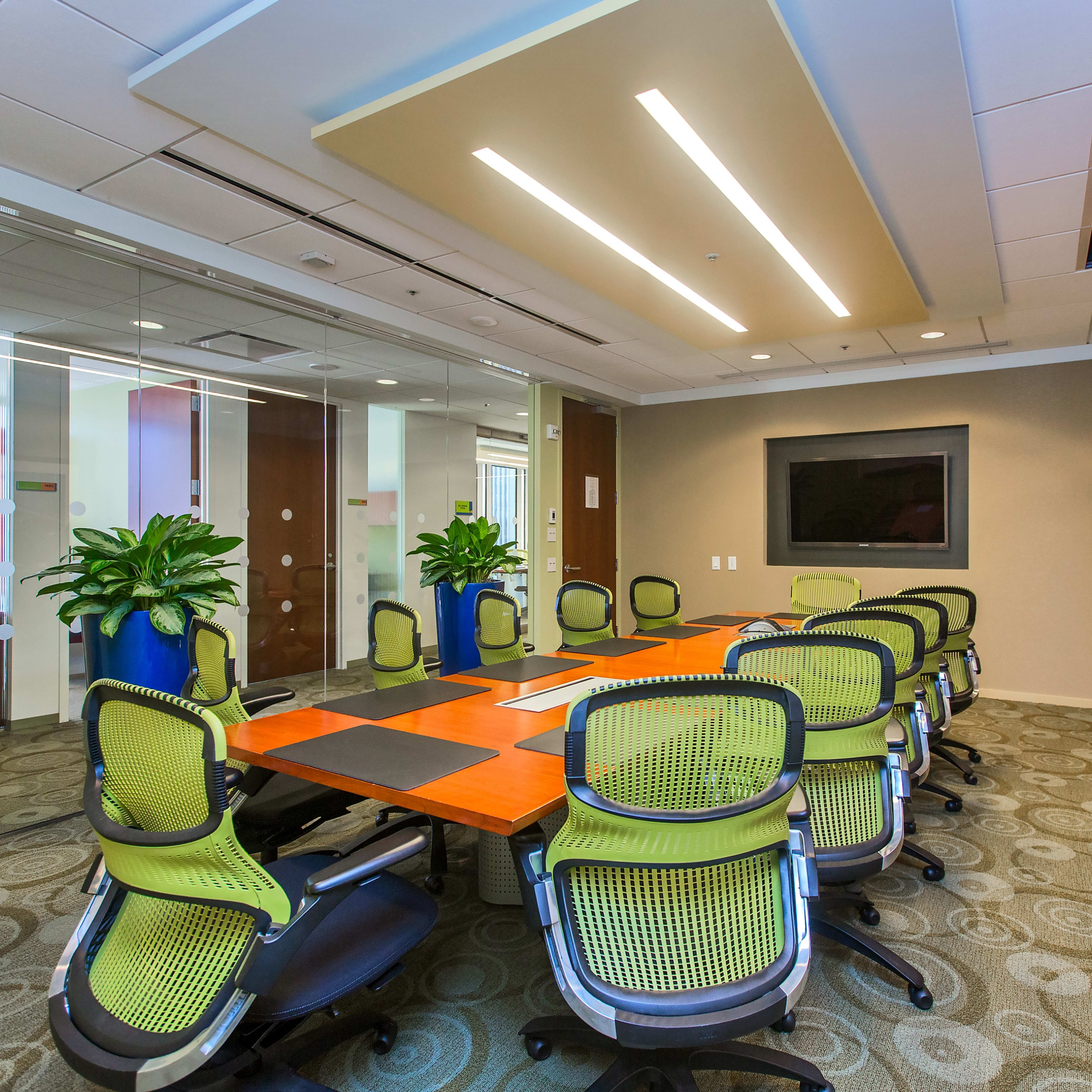 A modern conference room with a large orange table surrounded by green office chairs and plants, featuring glass walls and a mounted screen.