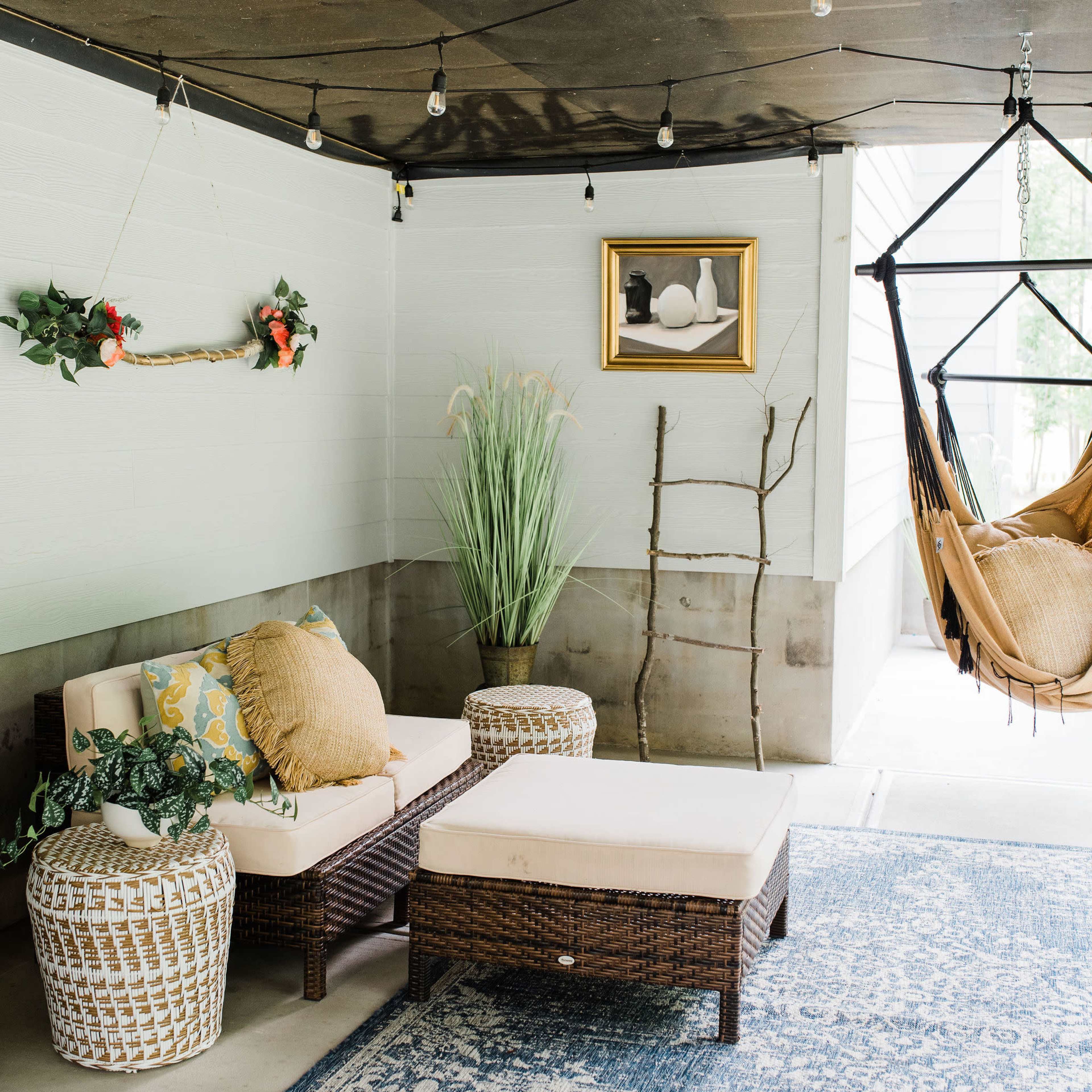 A cozy outdoor seating area with a wicker couch, an ottoman, a hanging chair, and decorative plants against a light blue wall.