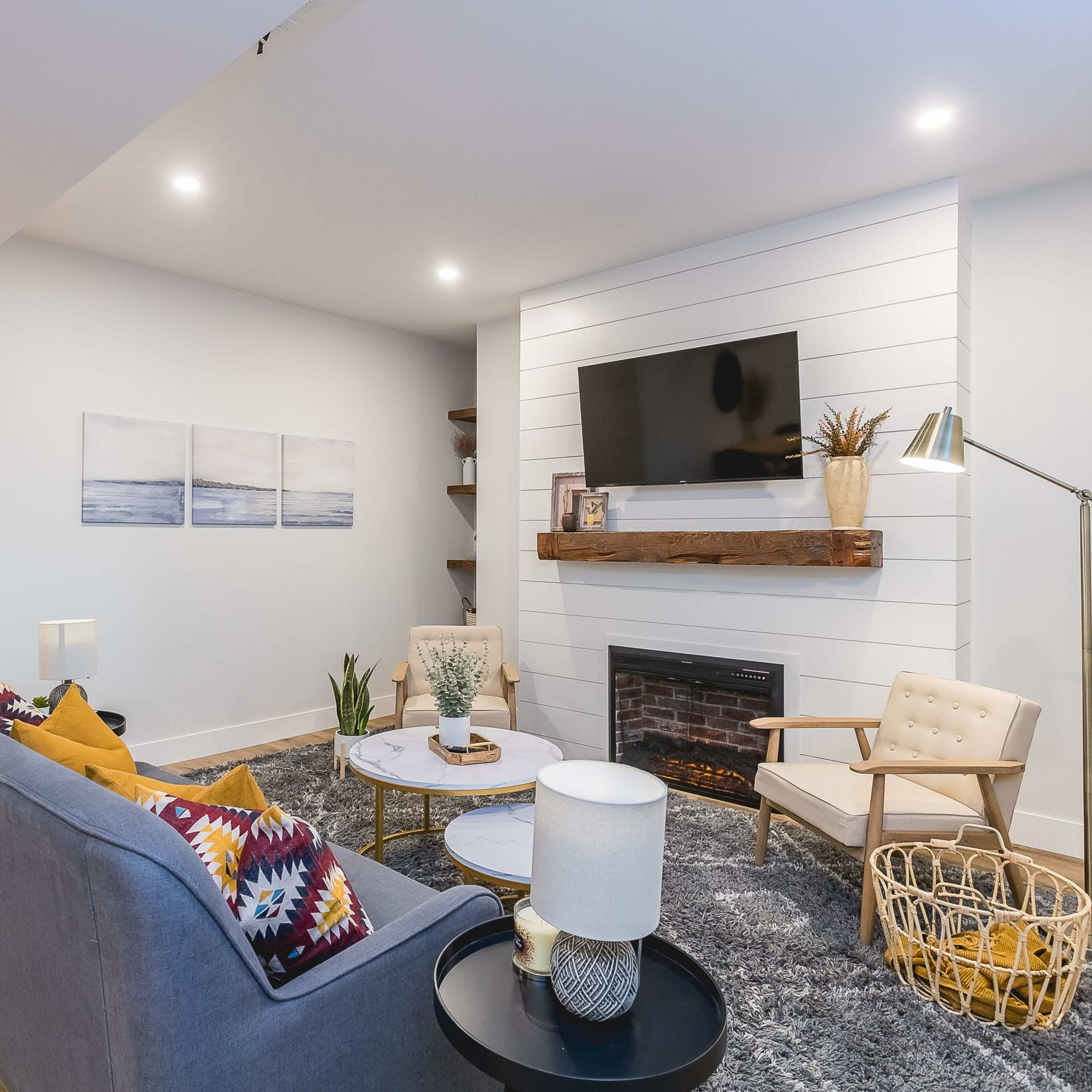 A modern living room with a gray sofa, a coffee table, a mounted television above a white shiplap fireplace, and a cozy reading chair.