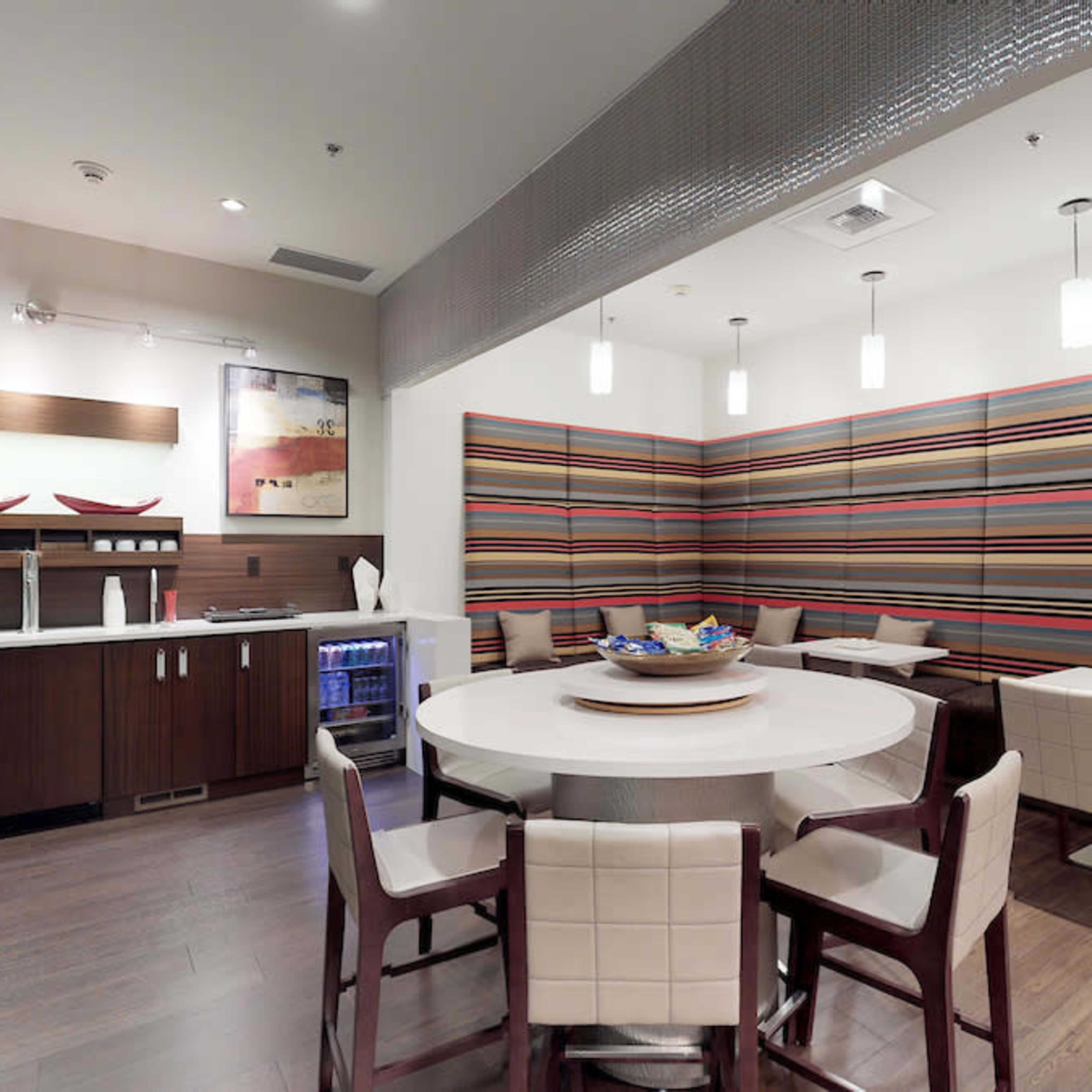 A modern dining area with a round table, six chairs, and a striped wall design, featuring a small kitchenette and a variety of decorative fruit on the table.