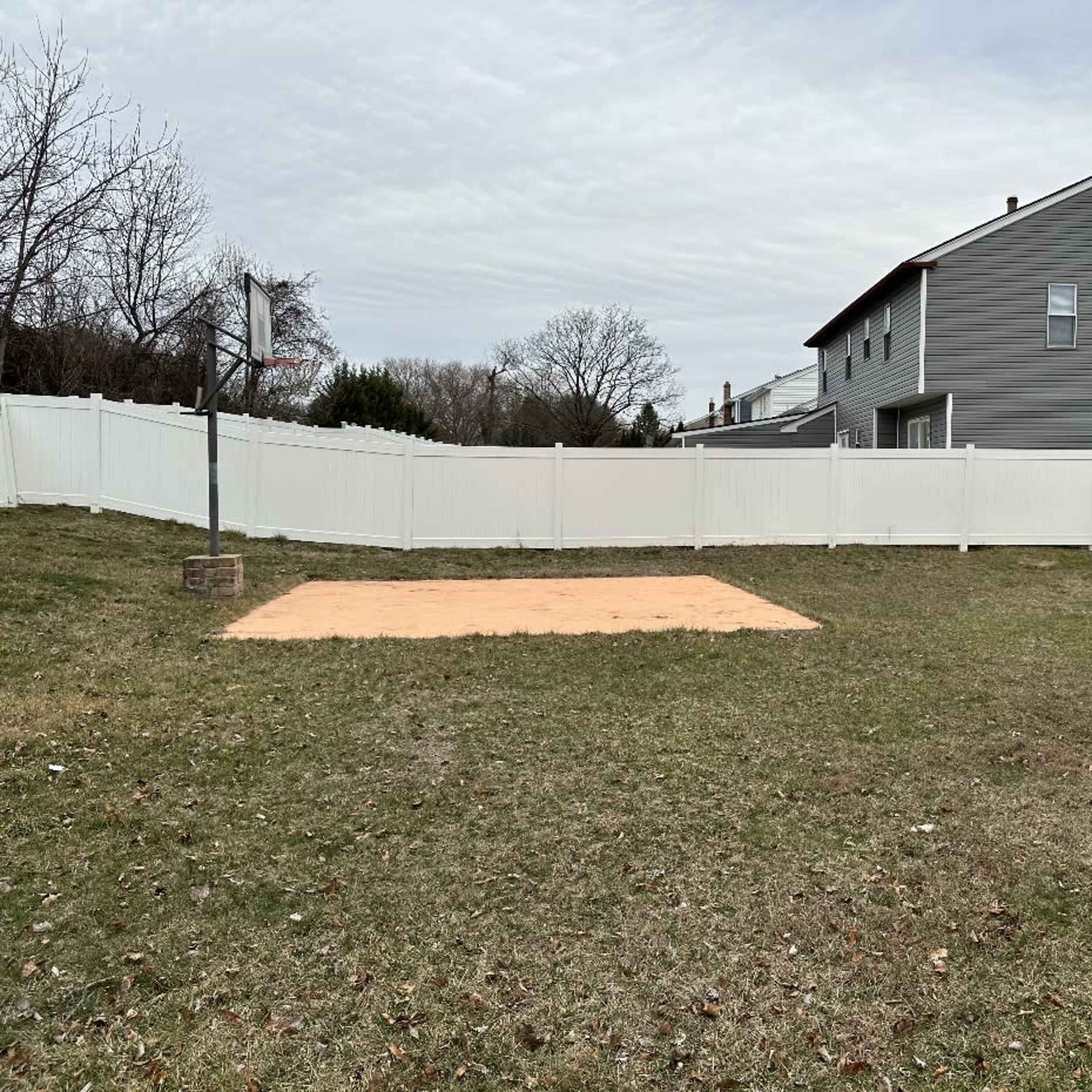 A flat, sandy area intended for a basketball court, situated in a backyard surrounded by a white fence and two buildings.