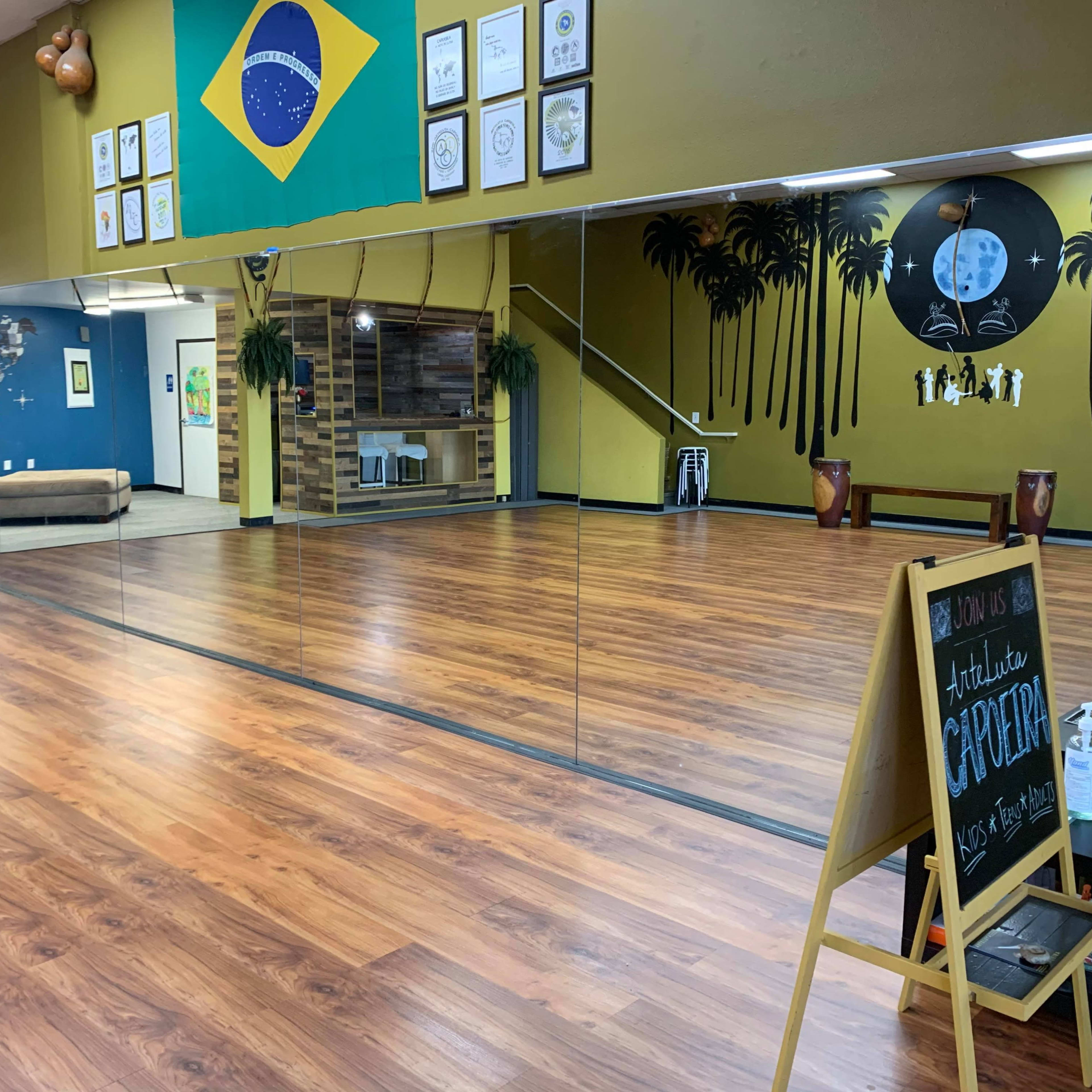 A spacious dance studio with wooden flooring, mirrors on one wall, a Brazilian flag hanging, and decorative elements like palm trees and a chalkboard sign.