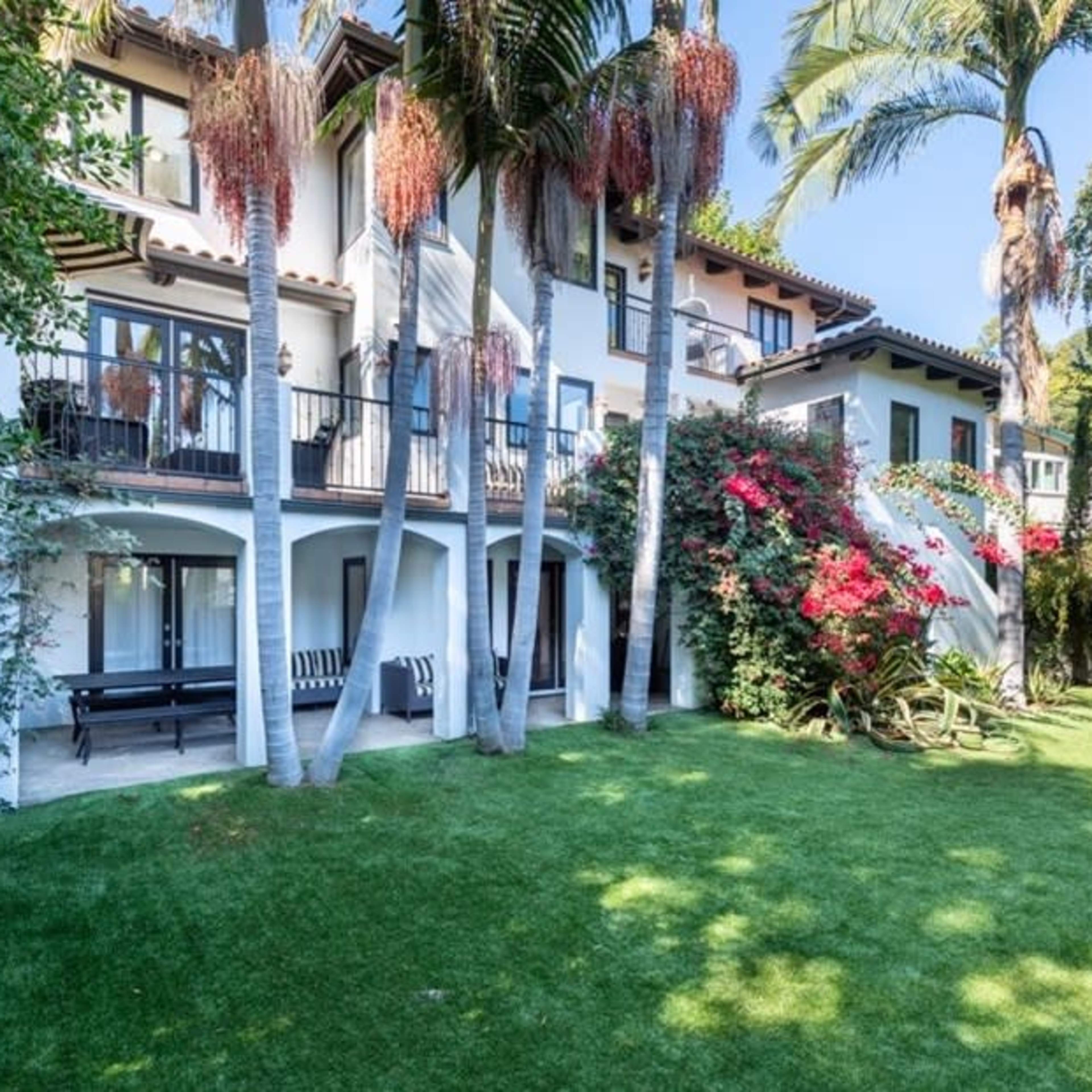 A multi-story house surrounded by palm trees and colorful flowering plants in a well-manicured lawn.