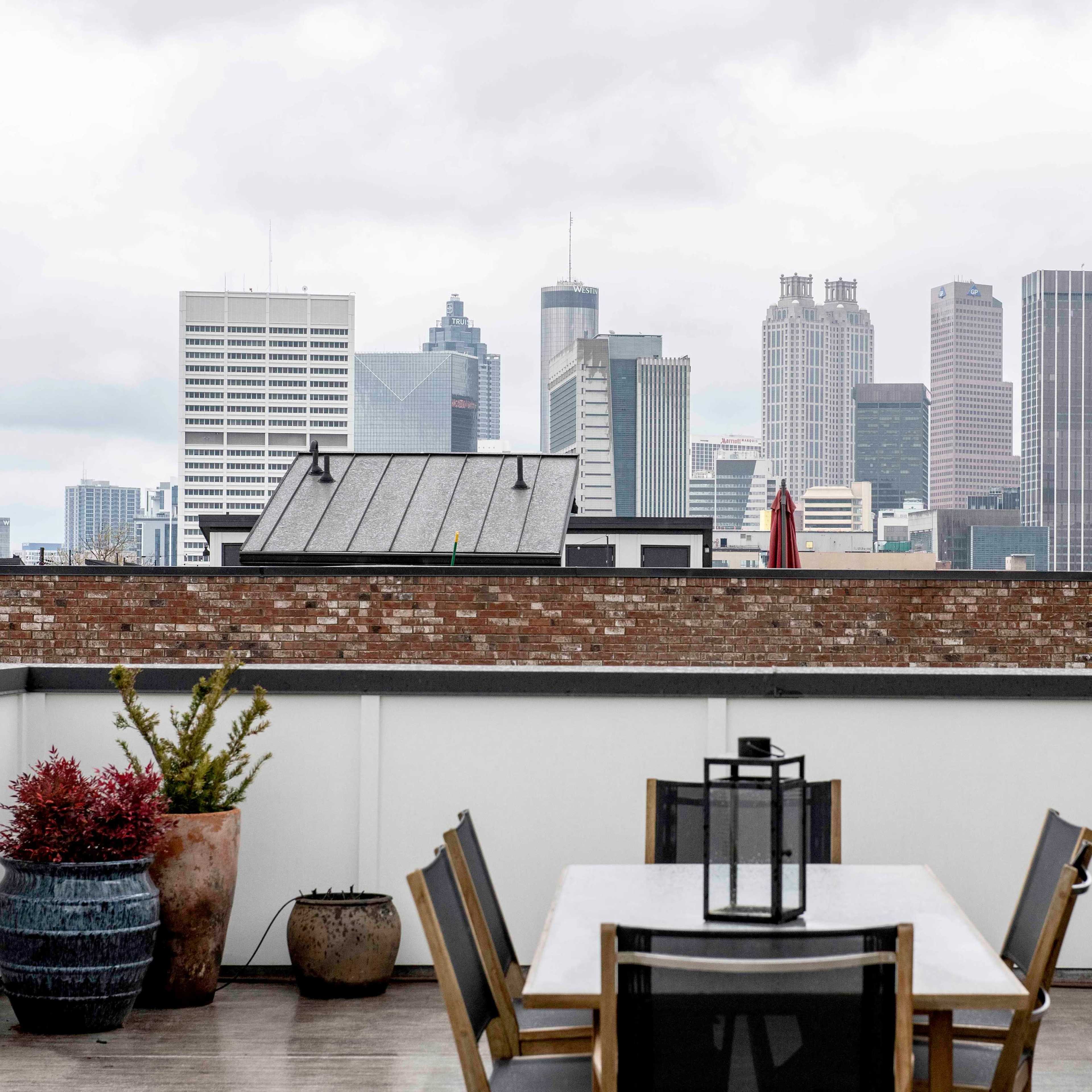 A rooftop terrace features a dining table and chairs, overlooking a city skyline with tall buildings under a cloudy sky.