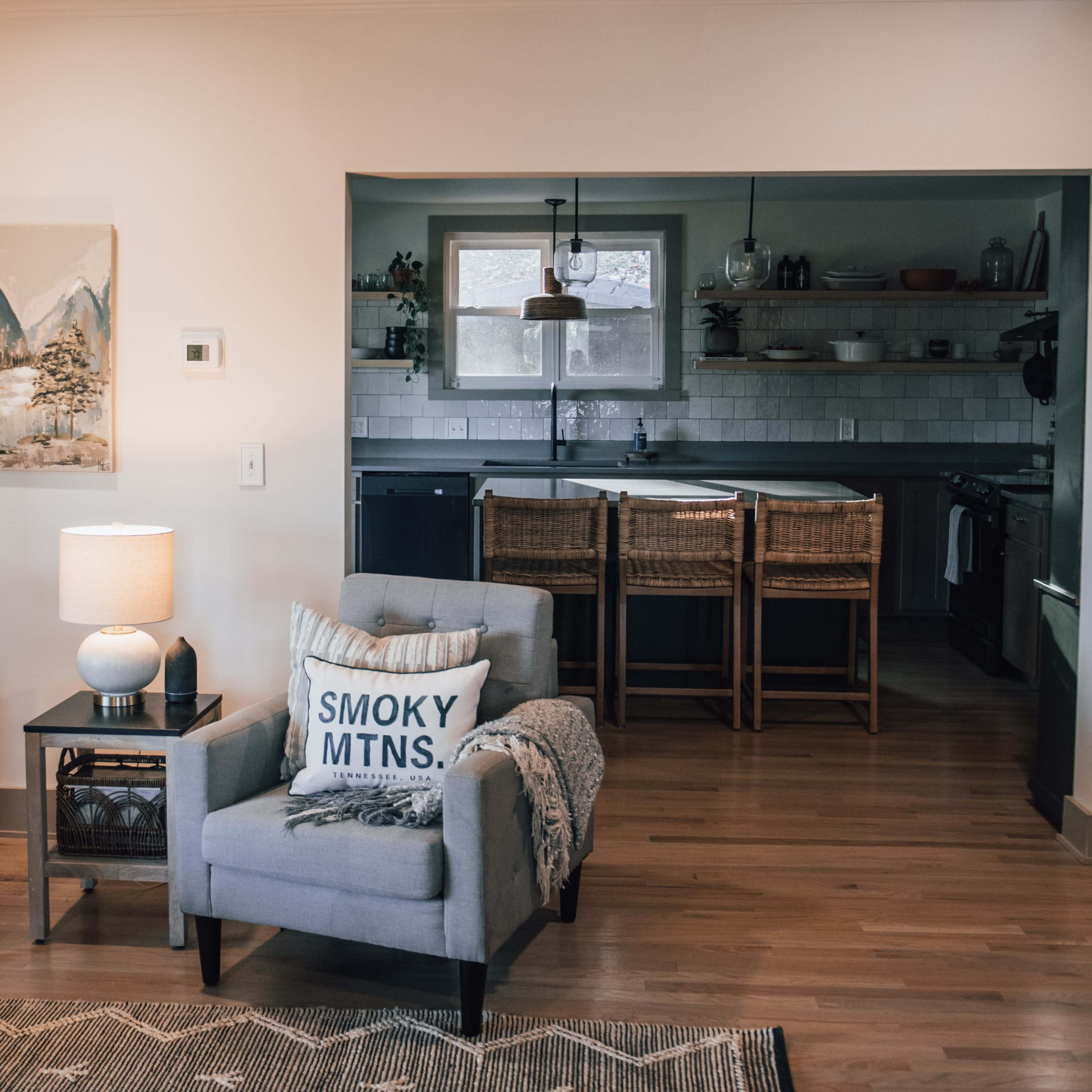 A cozy living room with two armchairs, a lamp, and a painting on the wall, leading to an open kitchen with wooden shelves and bar stools.