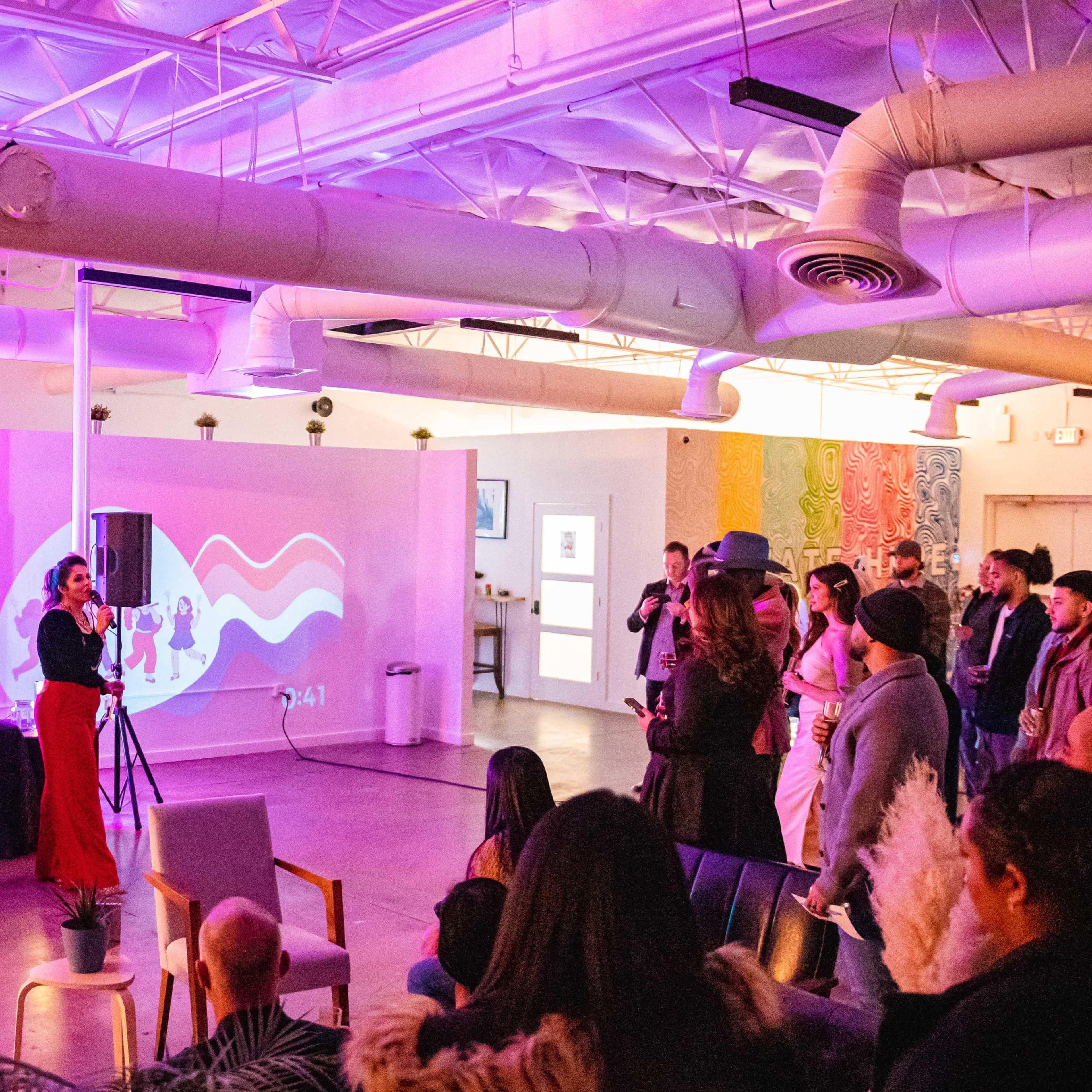A group of people stands in front of an audience in a brightly lit room, with a colorful projection on the wall behind them.