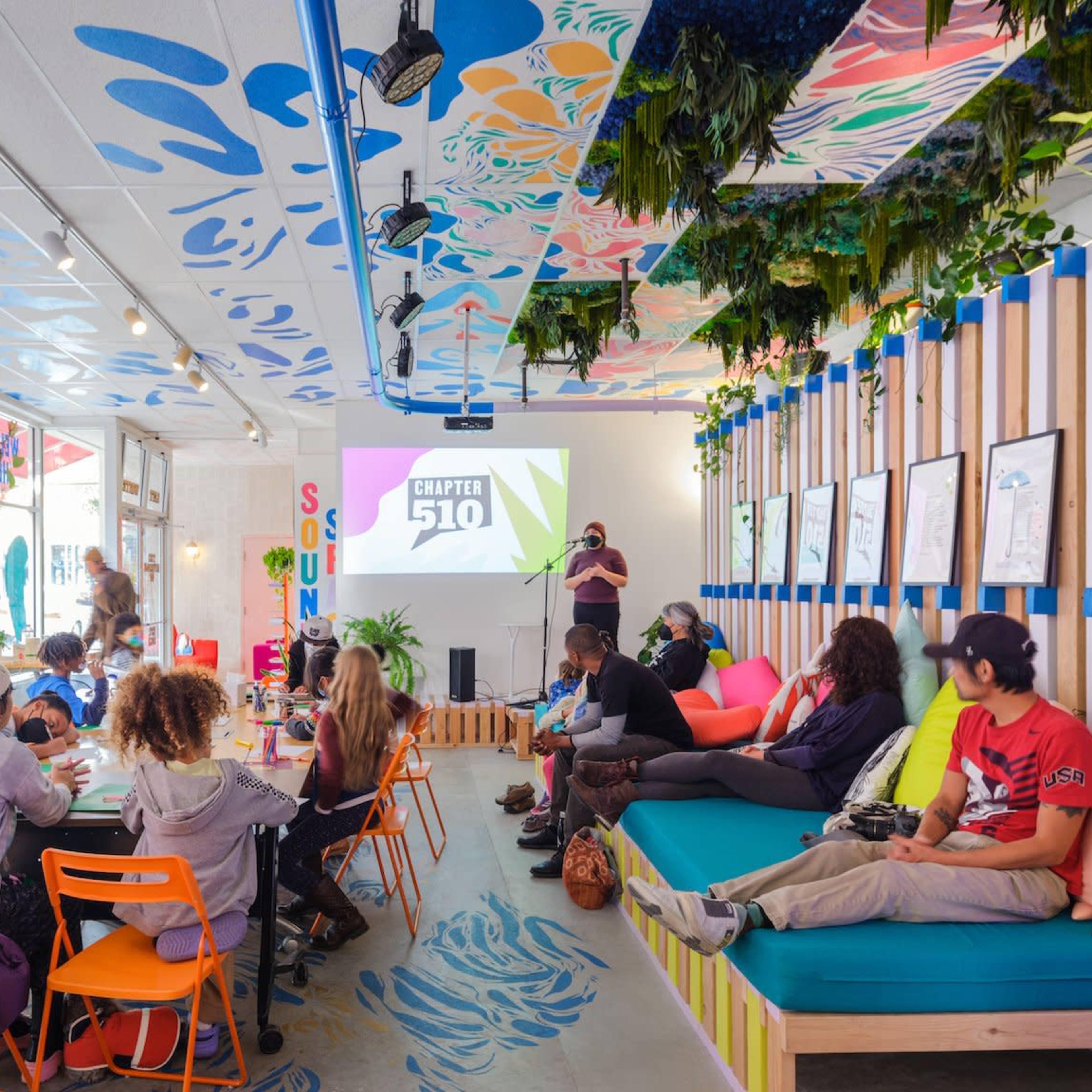 A lively café features colorful seating and a projector screen displaying "Chapter 510," while people engage in conversation and activities.
