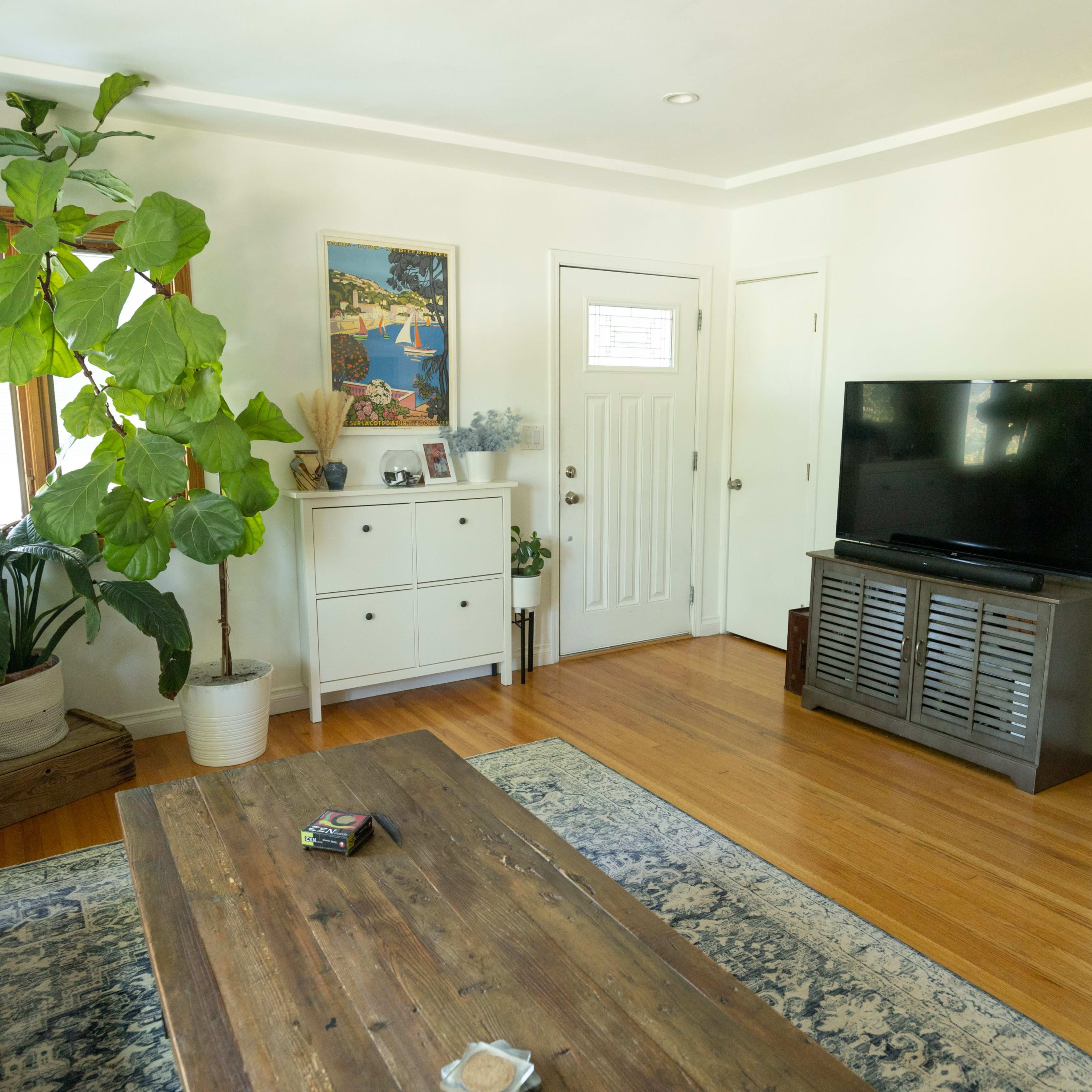 A living room features a sofa, a wooden coffee table, a sideboard, a television, and potted plants near a door and window.