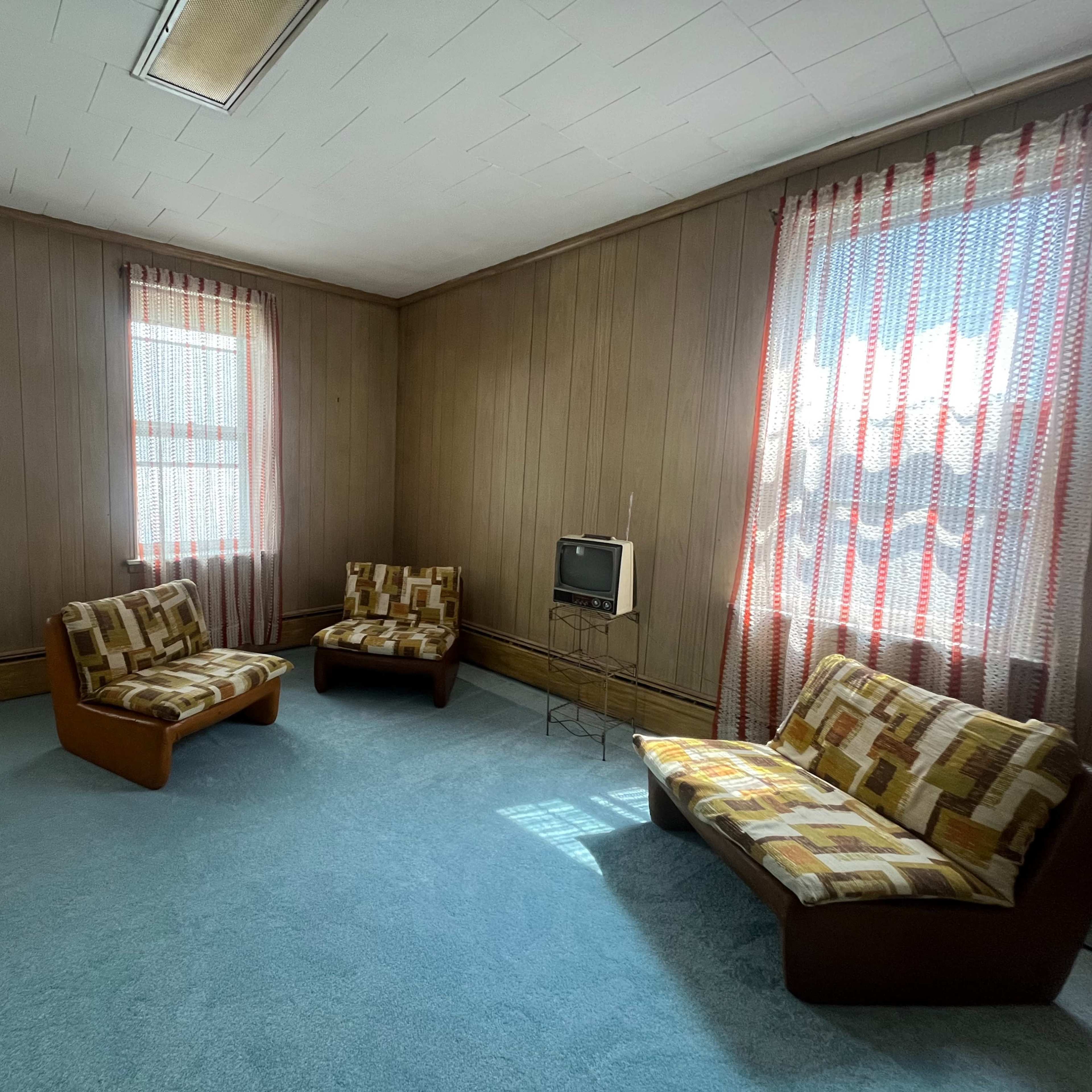 A sparsely furnished room with two patterned couches, a small television, and large windows covered by sheer curtains.