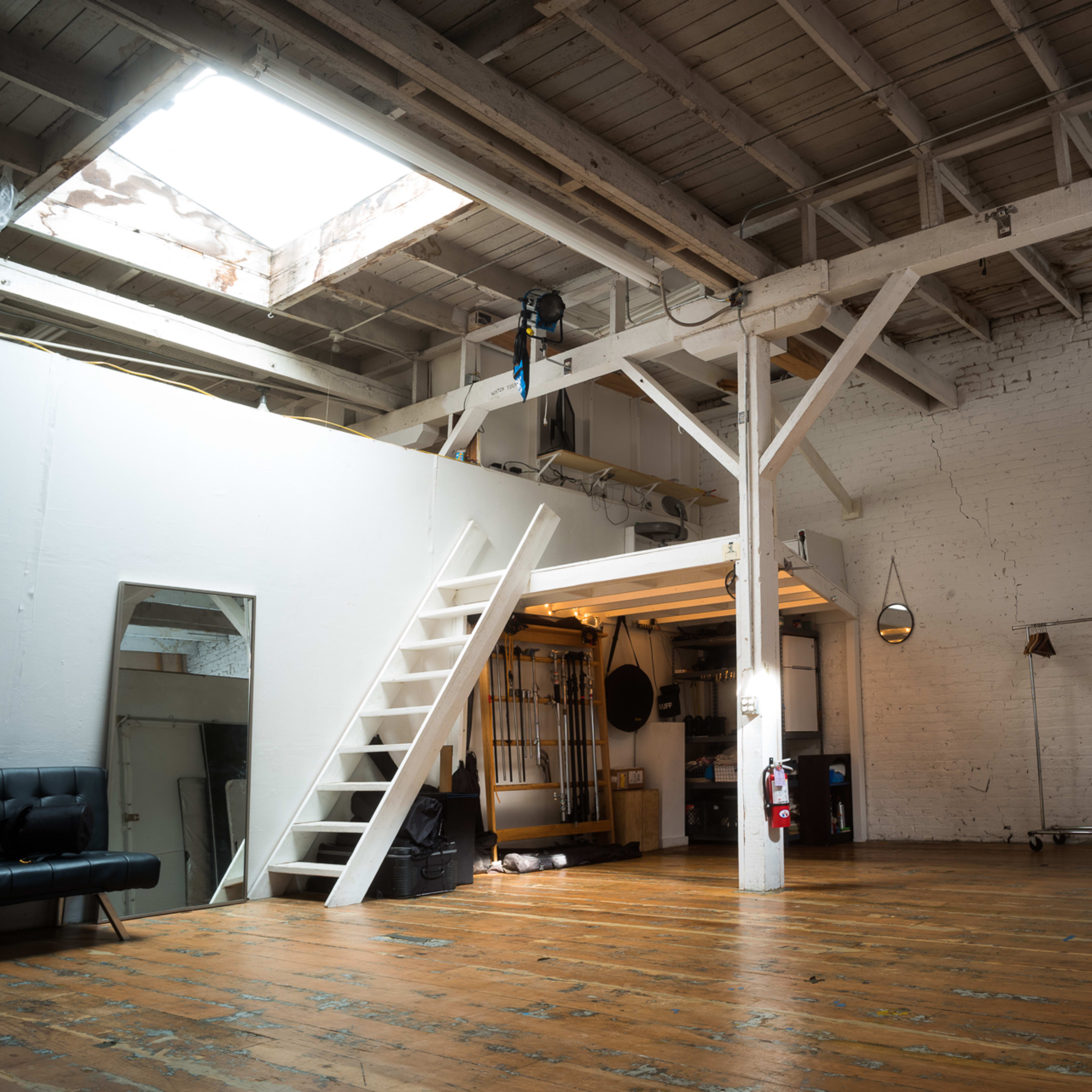 An open, spacious studio with wooden floors, exposed beams, and a ladder leading to a higher platform.