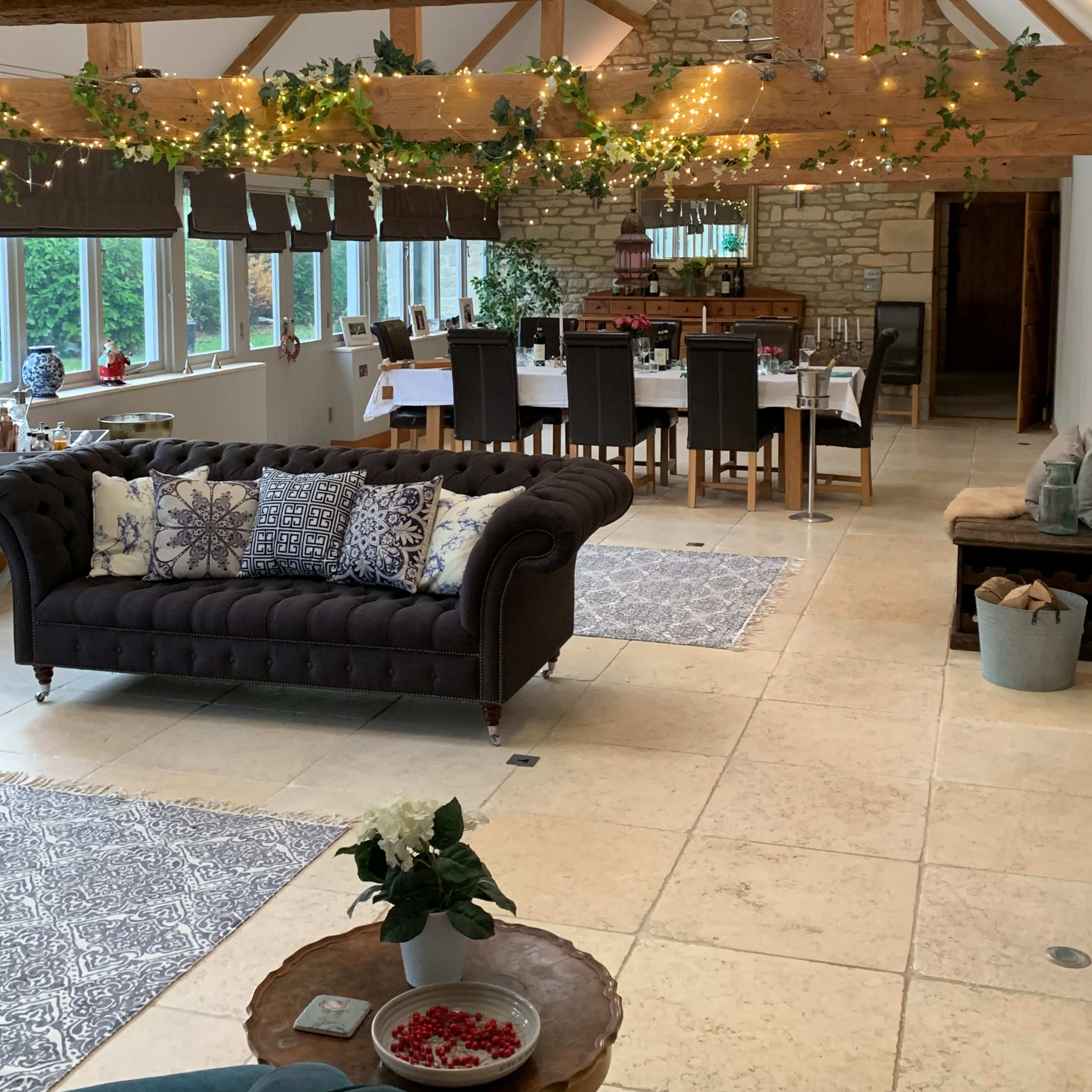 A spacious, open-plan room with a cozy seating area featuring a dark sofa, a dining table set for a meal, and wooden beams adorned with string lights.