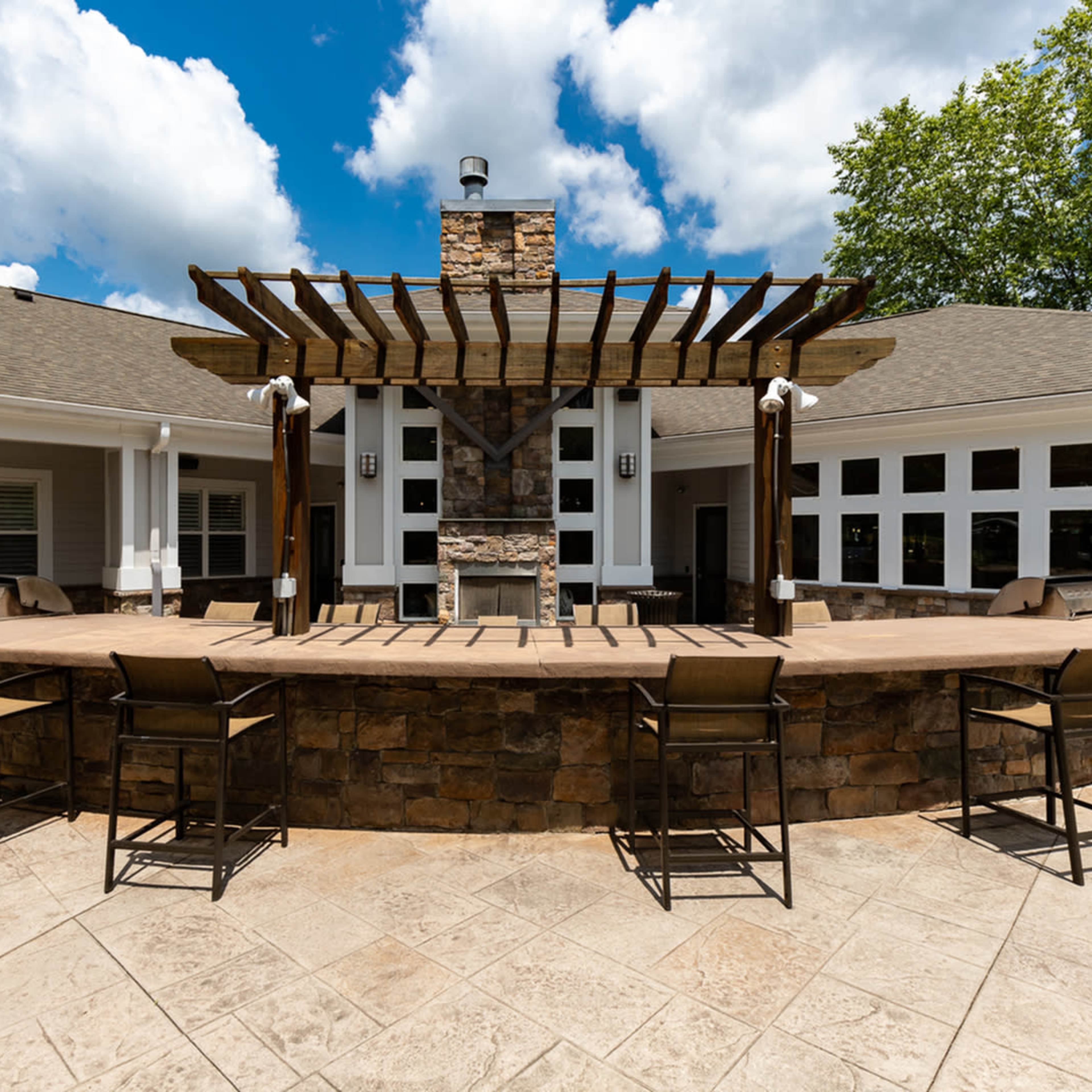 A patio area with a stone fireplace, a wooden pergola overhead, and bar stools arranged around a curved stone counter.