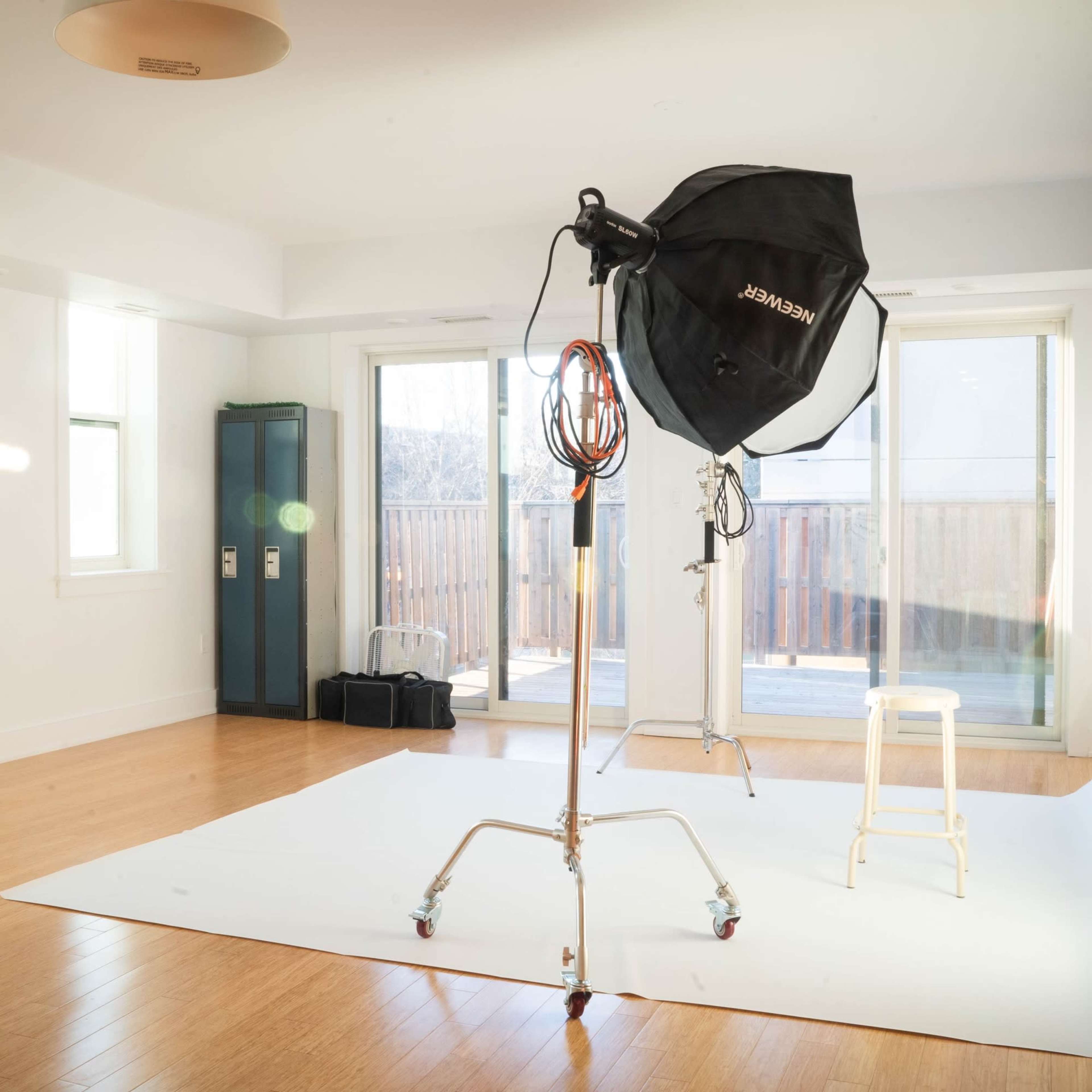 A photography studio features a white backdrop, a light stand with a softbox, and a stool, with large windows allowing natural light.